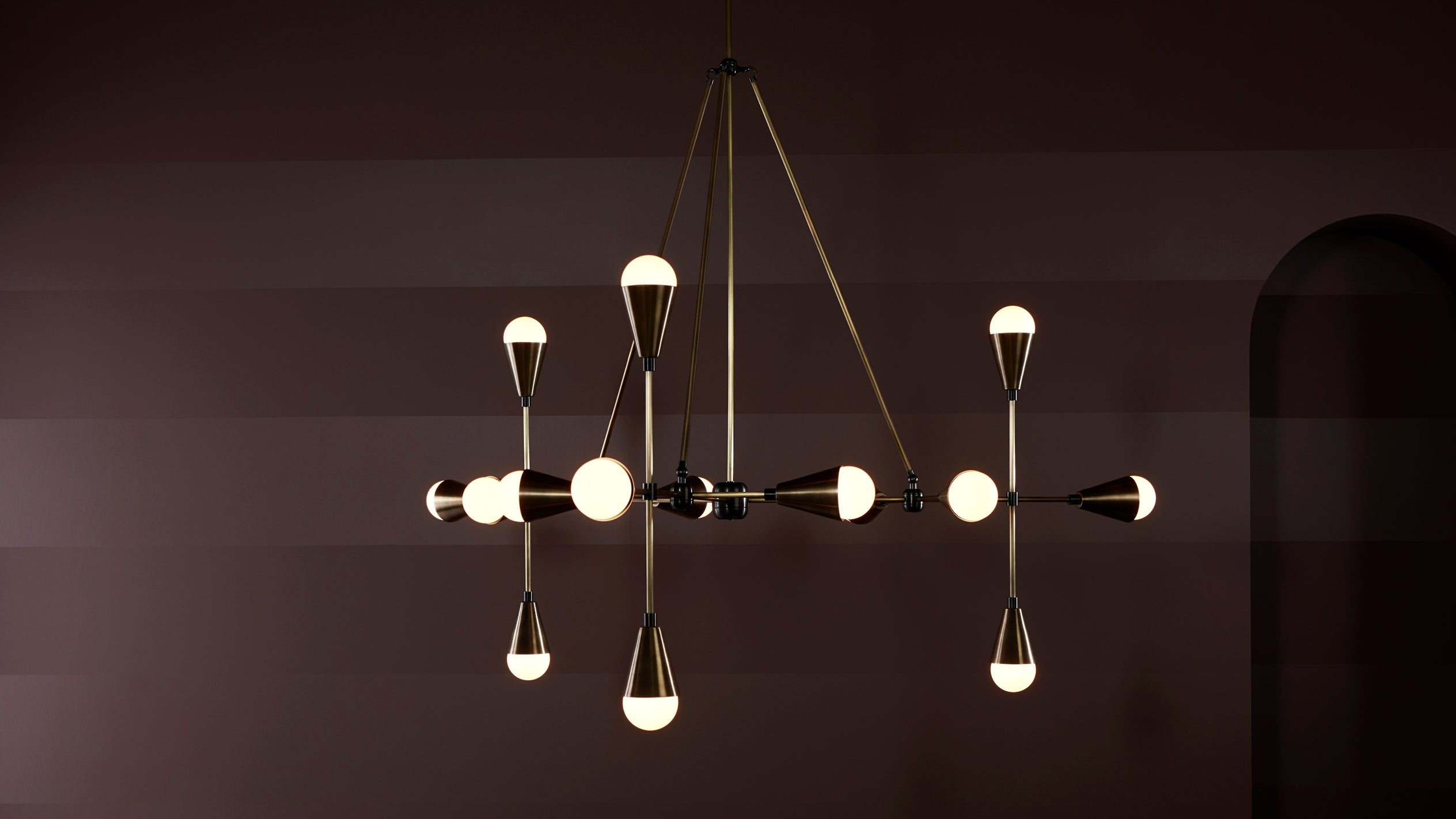 Modern black and brass chandelier with multiple illuminated conical bulbs suspended against a dark striped wall