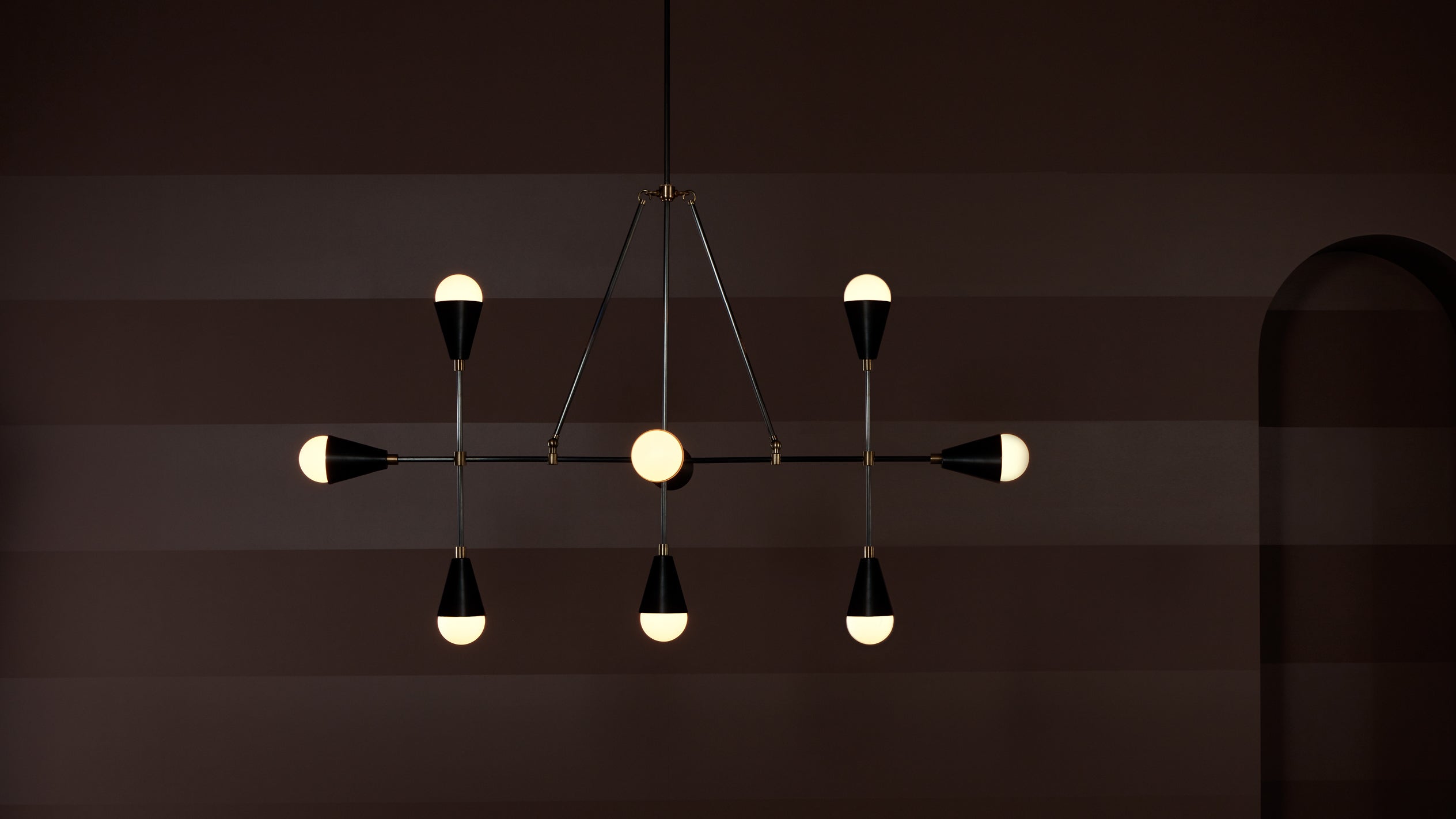 Modern chandelier with seven teardrop-shaped light bulbs suspended from a minimal black metal frame against a dark brown wall