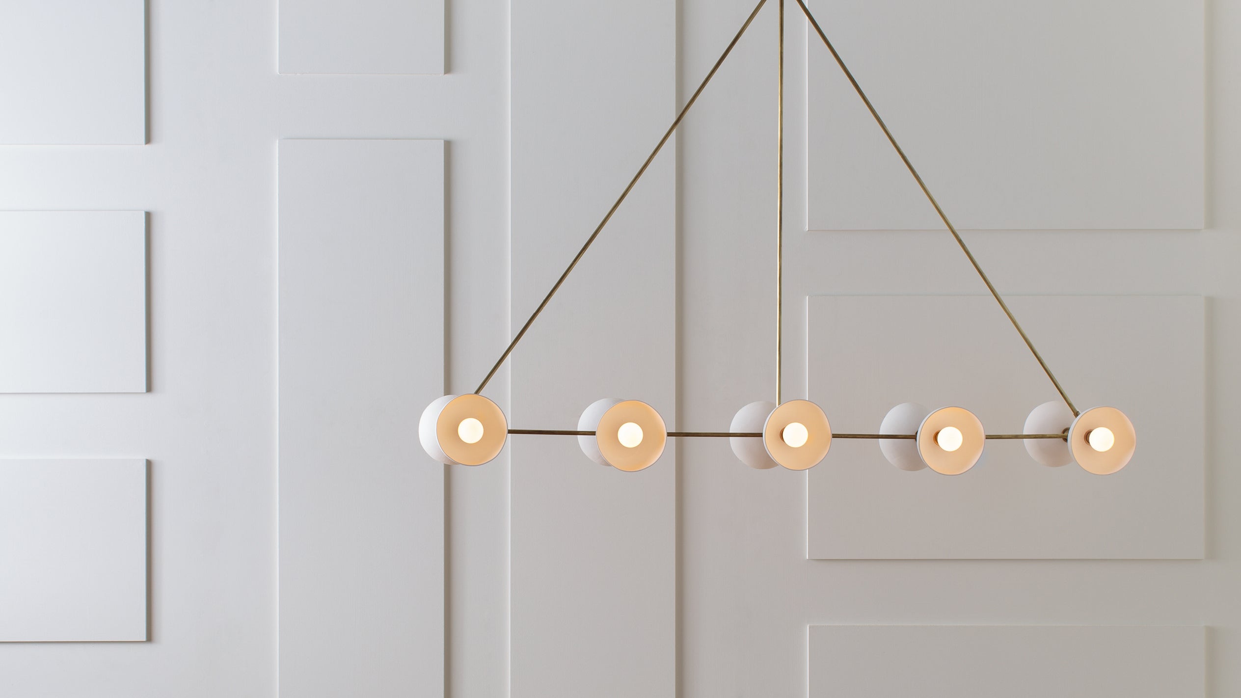 Minimalist linear chandelier with six round white lampshades suspended against a white paneled wall background