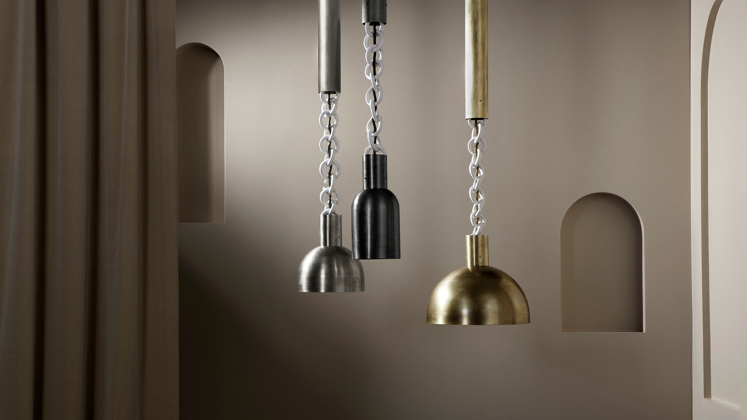 Three hanging metal pendant lights in silver, black, and brass finishes against a beige wall with arched niches