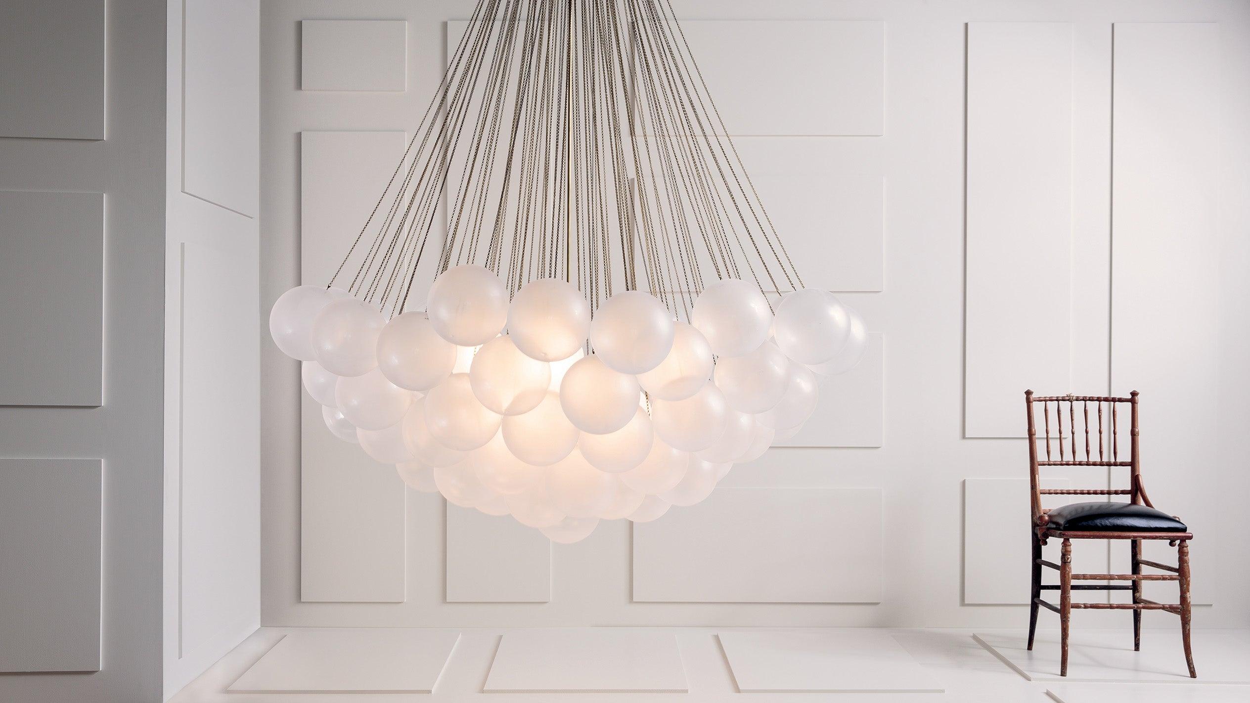 Cluster of frosted glass globe pendant lights suspended by thin cords in a white paneled room with a dark wooden chair