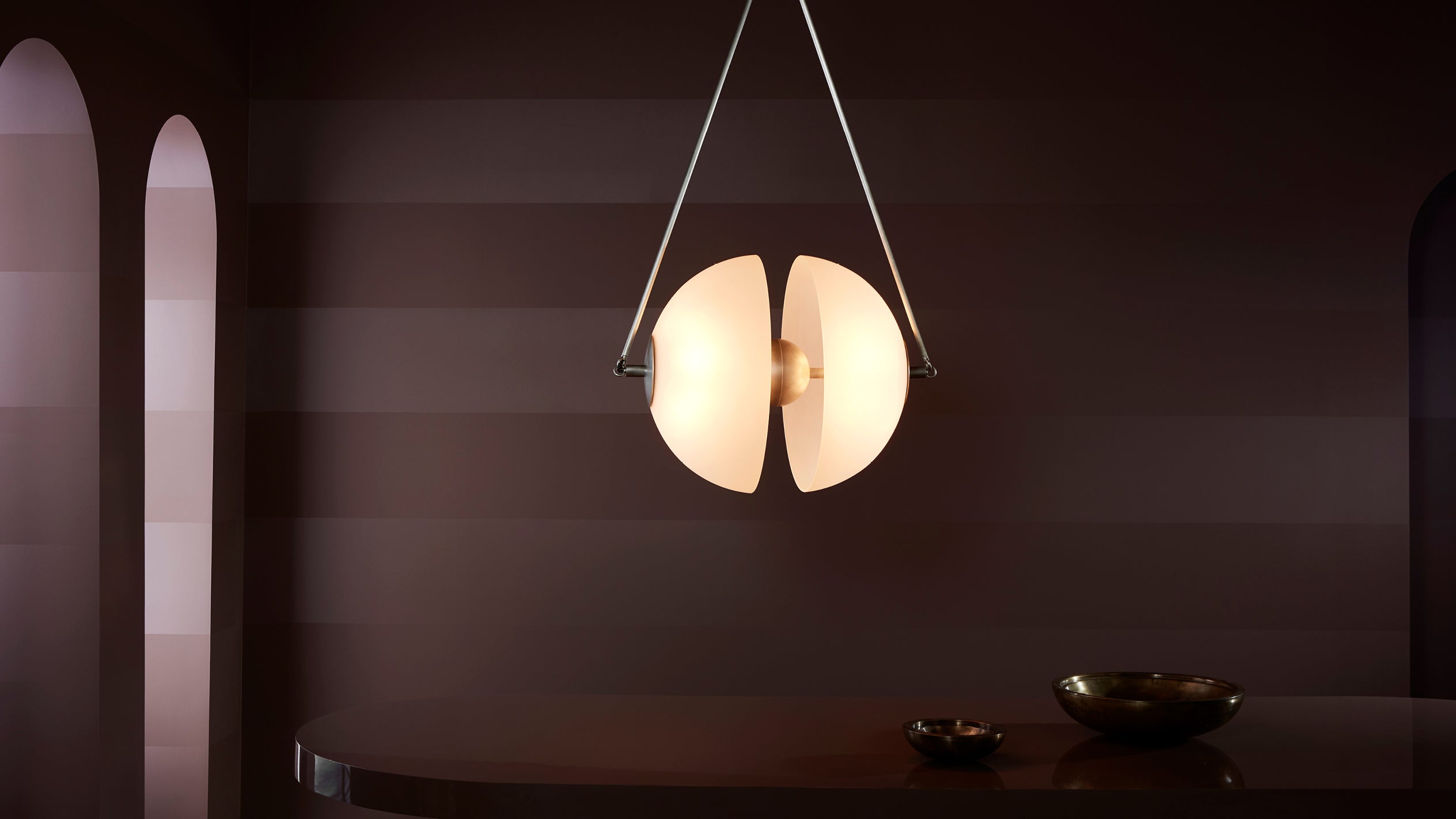 Modern pendant light with two curved frosted glass panels hanging above a dark wooden table in a muted room.