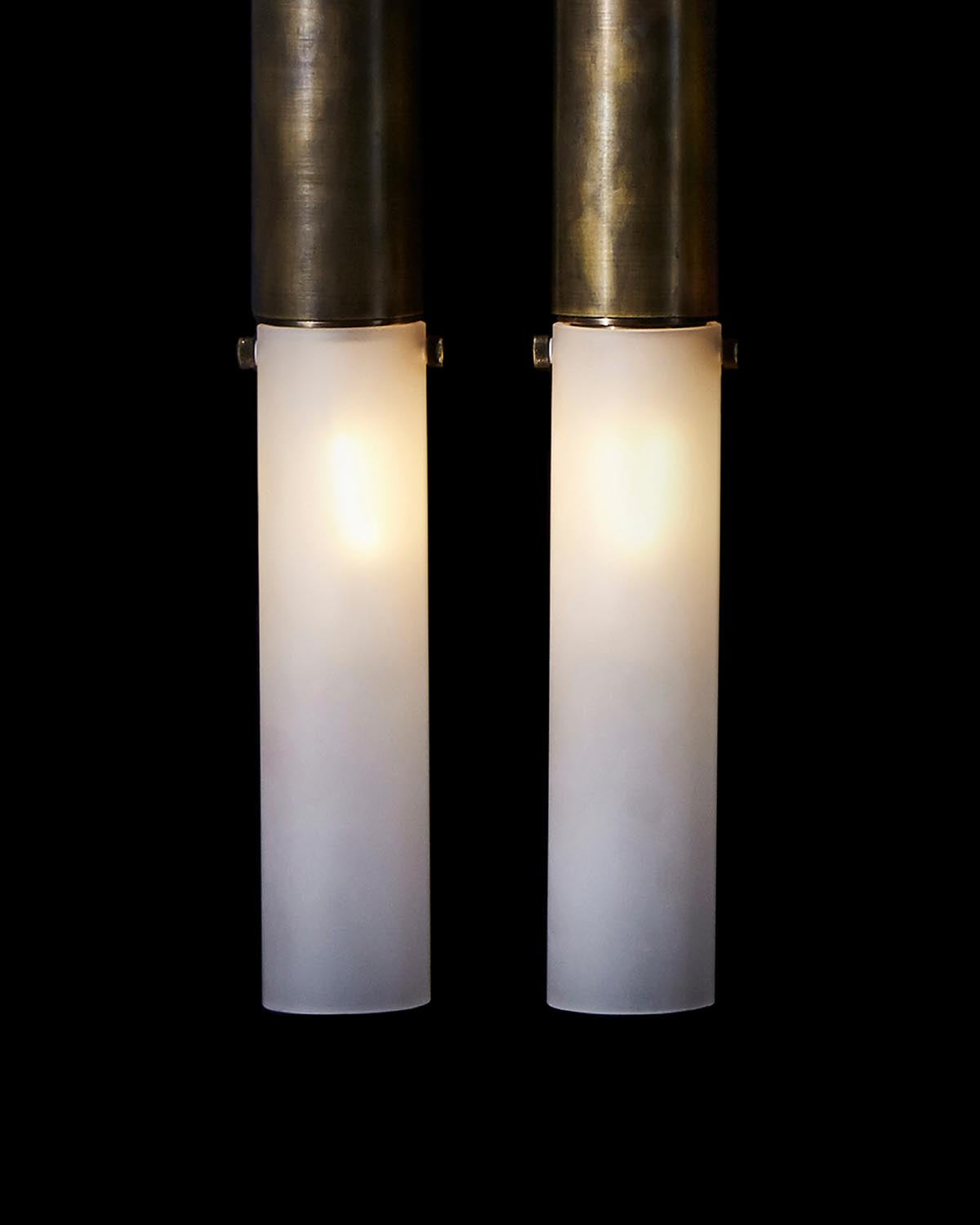 Pair of vertical wall sconces with frosted cylindrical glass shades and aged brass fittings against black background
