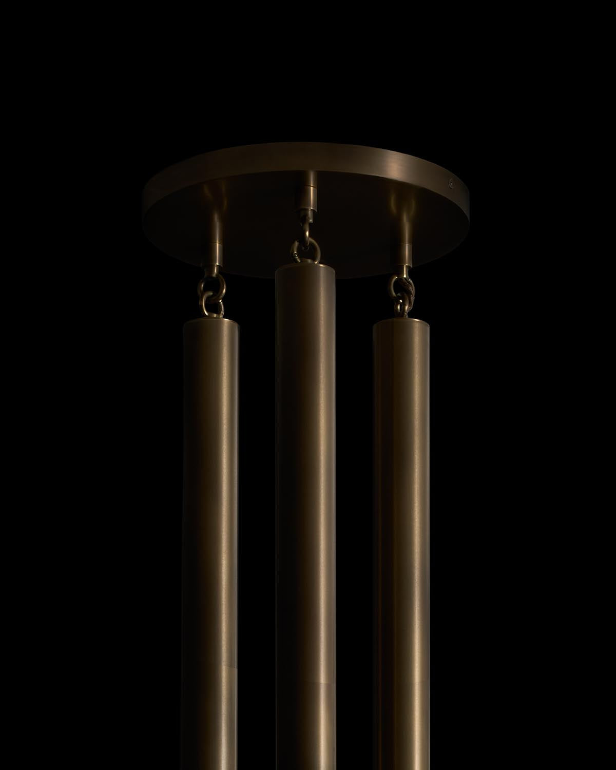 Close-up of three long cylindrical bronze pendant rods hanging from a round bronze ceiling mount against black background