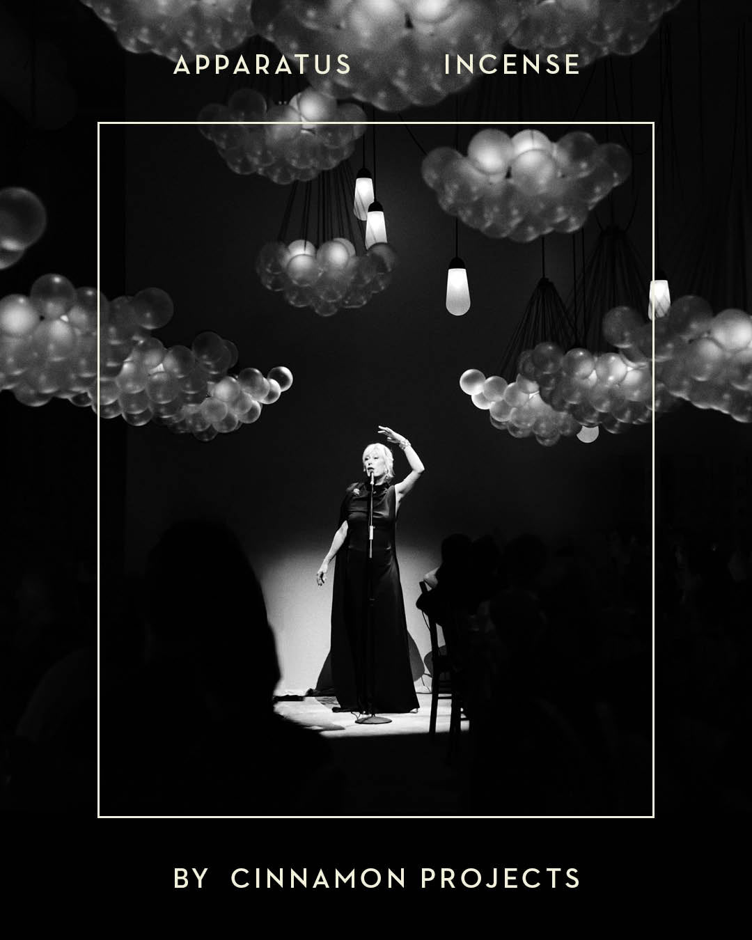 Black and white image of a singer on stage under cloud-shaped pendant lights with audience silhouettes in foreground