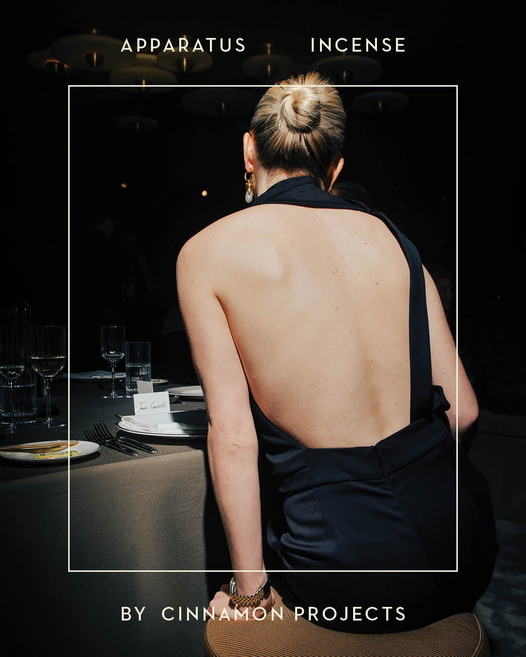 Woman in a black backless dress seated at a dining table set with glassware and plates in a dimly lit room