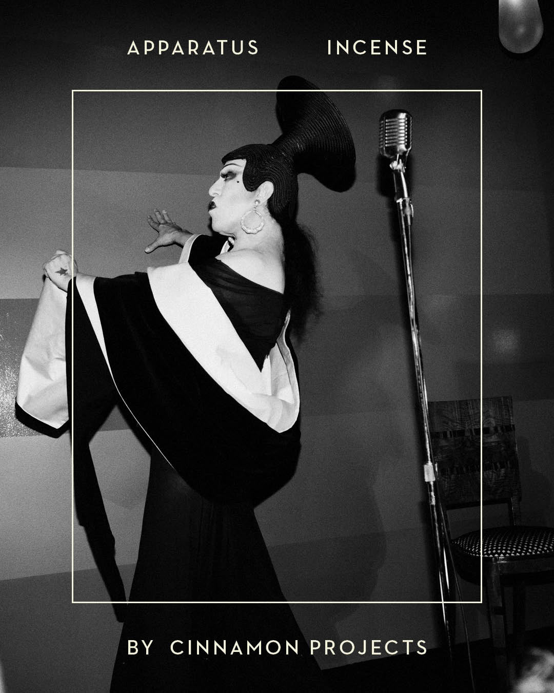 Black and white image of a performer in dramatic costume and headpiece beside a vintage microphone and chair against a plain background