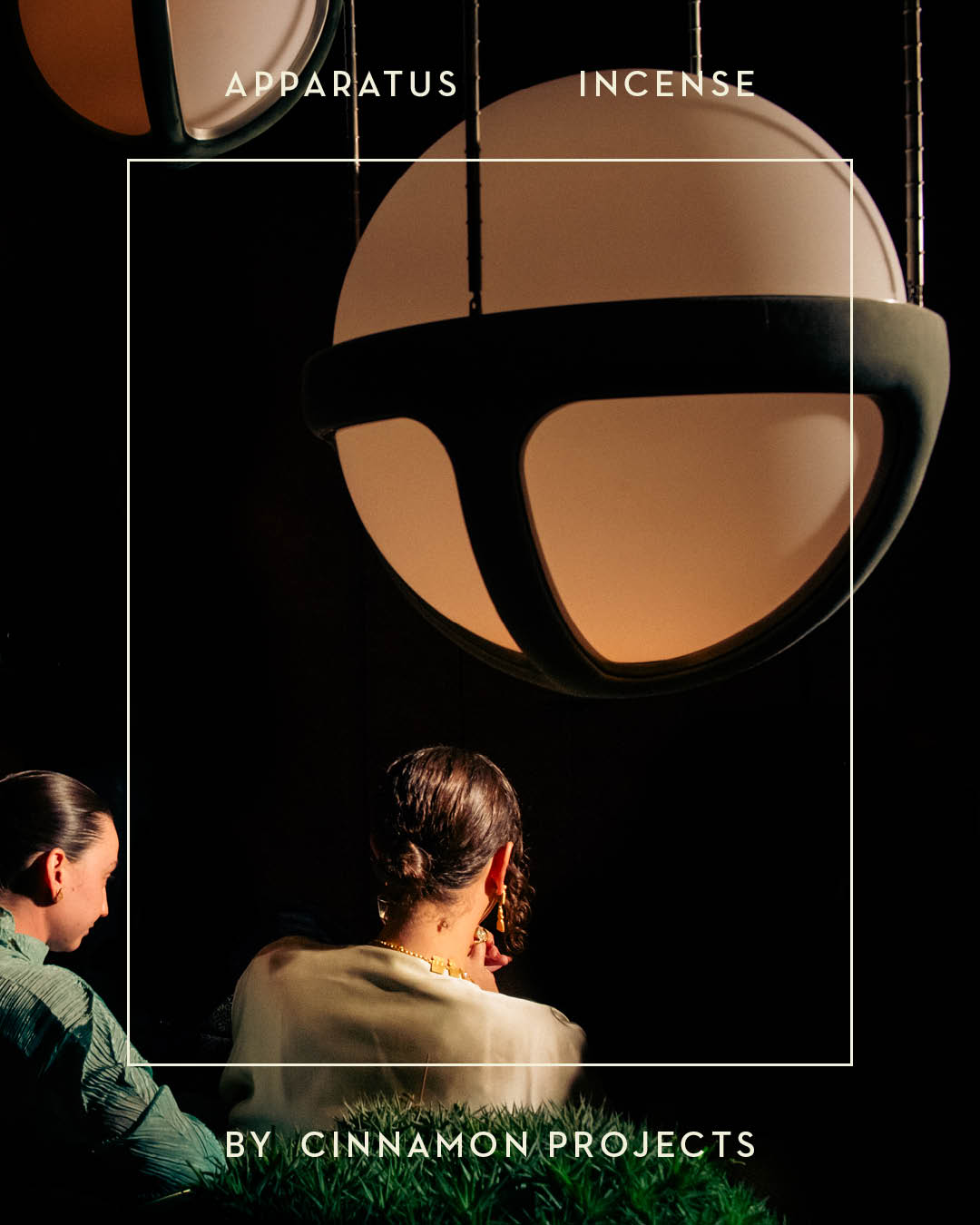 Two people seated under a large round pendant light with dark metal frame in a dim setting