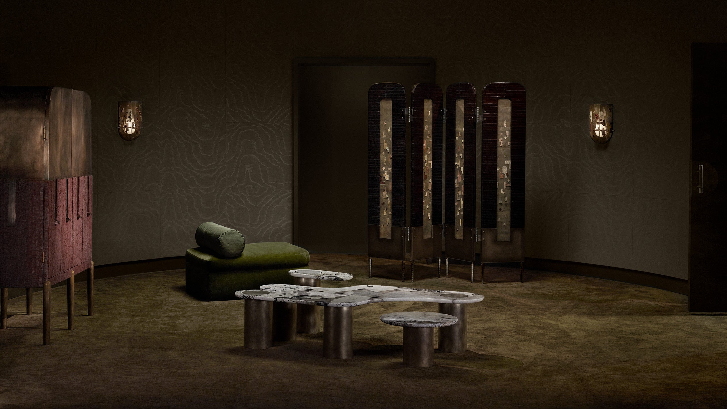 Dimly lit room with olive green carpet, sculptural coffee tables, a green lounge chair, and a folding screen against textured walls.