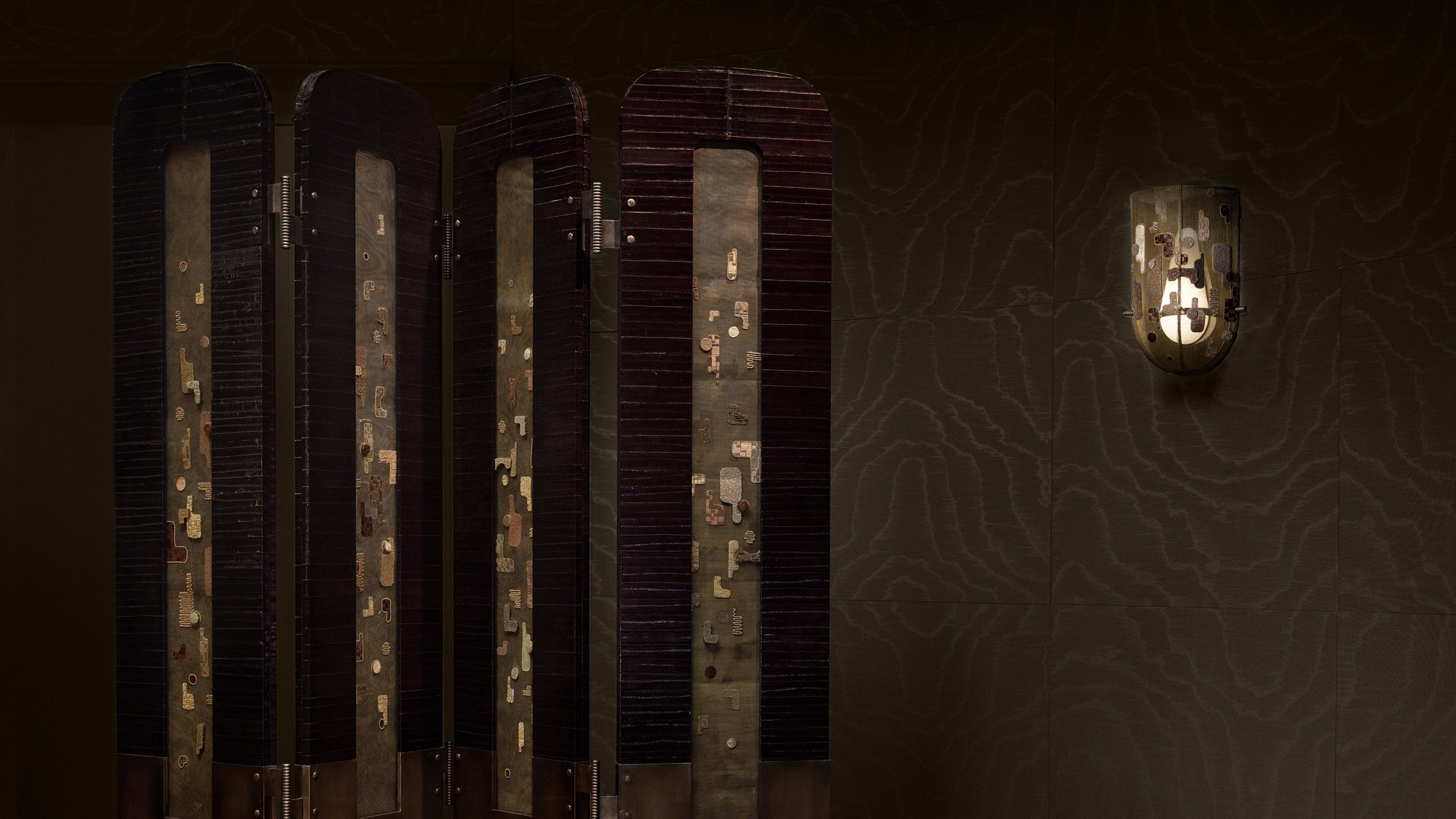 Dark wooden folding room divider with metallic gold rectangular accents next to a small wall-mounted light fixture on textured wall