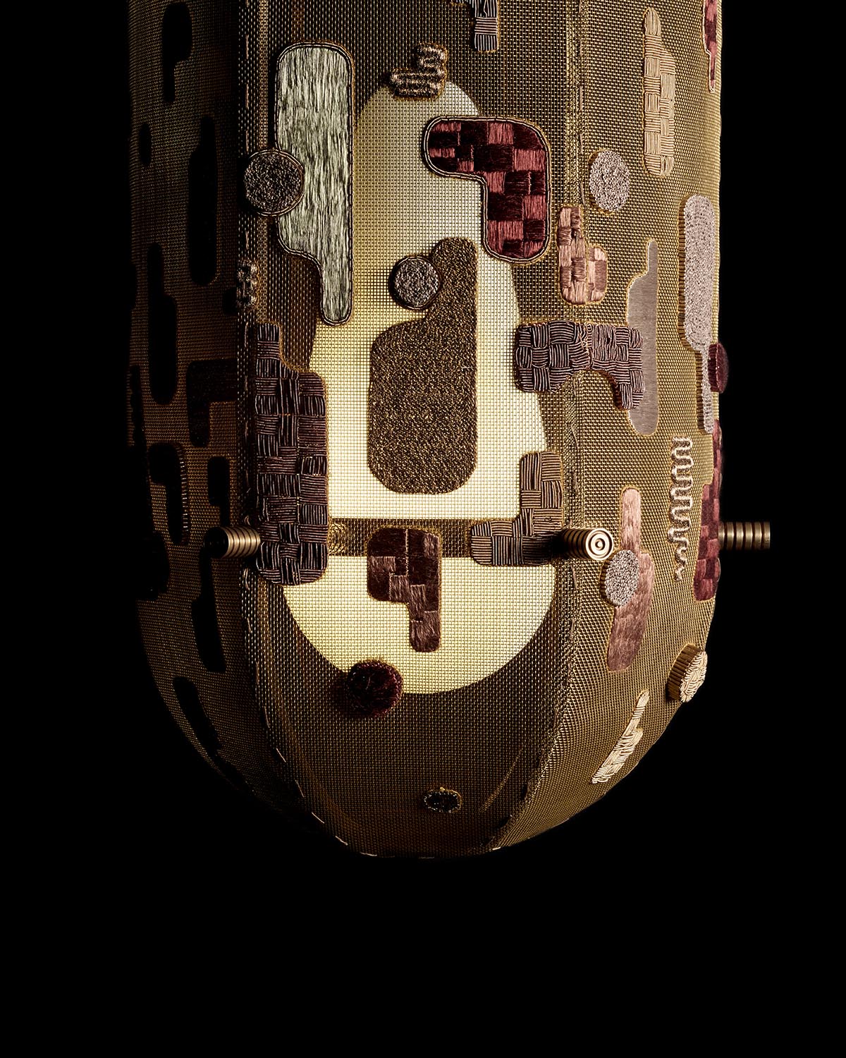 Close-up of hand-embroidered fabric covering an elongated sconce with abstract shapes in earthy tones against black background