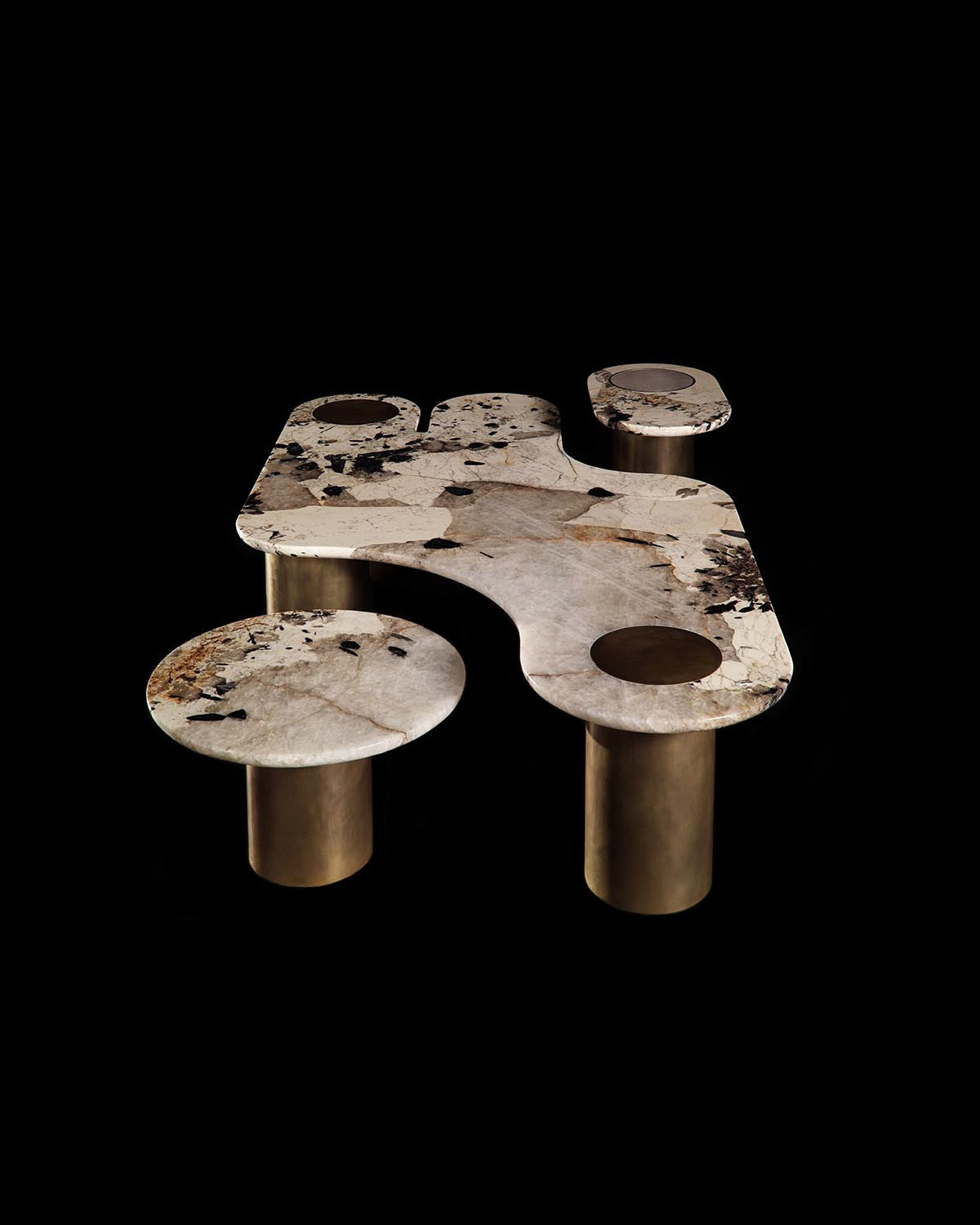 Asymmetrical marble table with mixed beige and black veining supported by cylindrical metallic bronze legs on black background