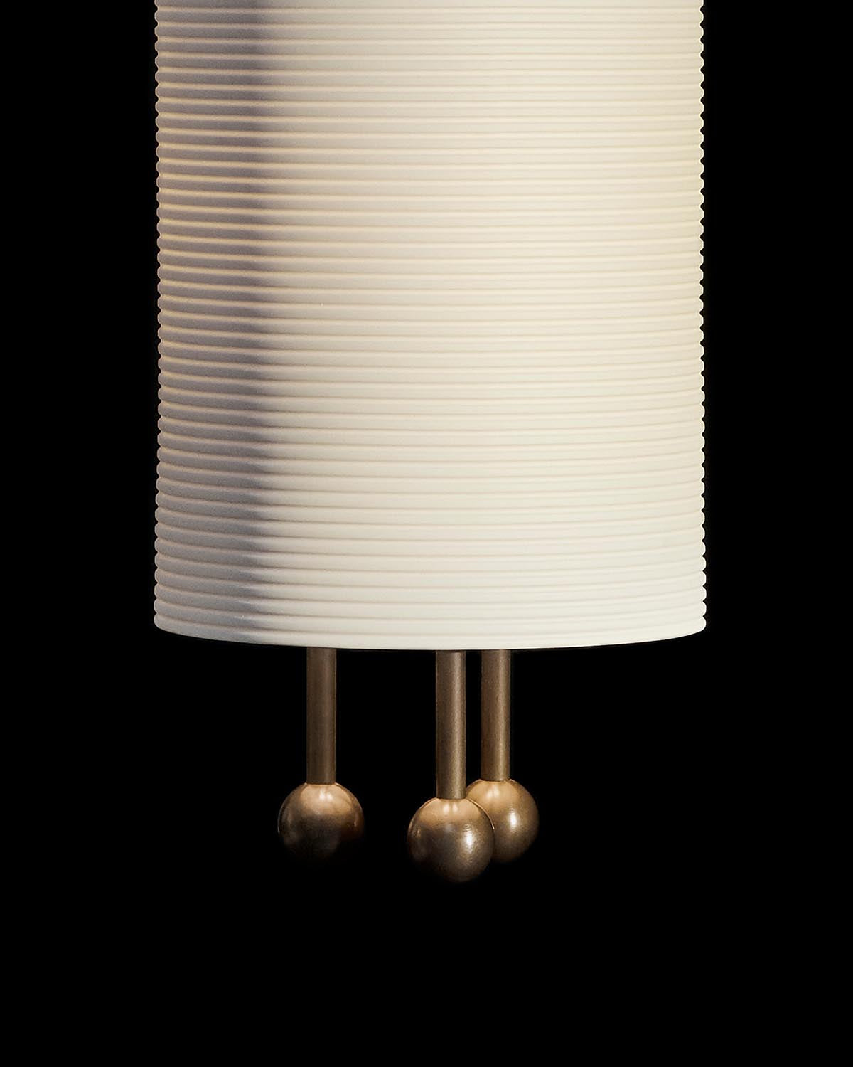 Cream ribbed cylindrical shade with three short brass rods ending in spherical tips against black background