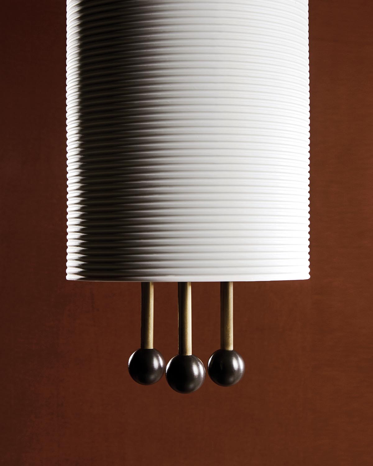 Close-up of a white ribbed cylindrical pendant lantern with three brass rods ending in dark spherical accents against a brown background