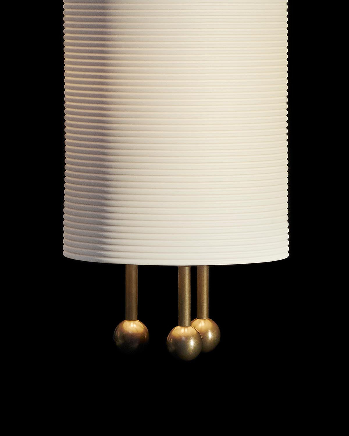 Close-up of a white ribbed cylindrical pendant lamp with three brass rods ending in round finials on a black background