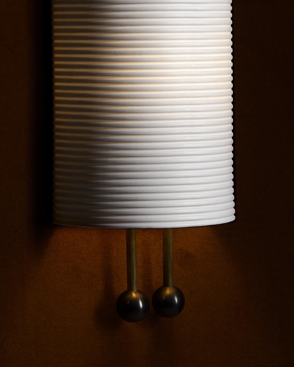 Close-up of a ribbed white cylindrical wall sconce with two brass pull chains against a brown background