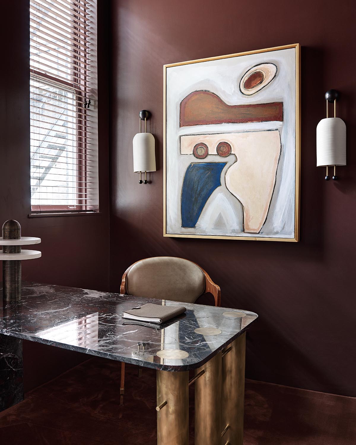 Pair of modern white cylindrical sconces mounted on dark brown wall beside abstract framed artwork above marble desk
