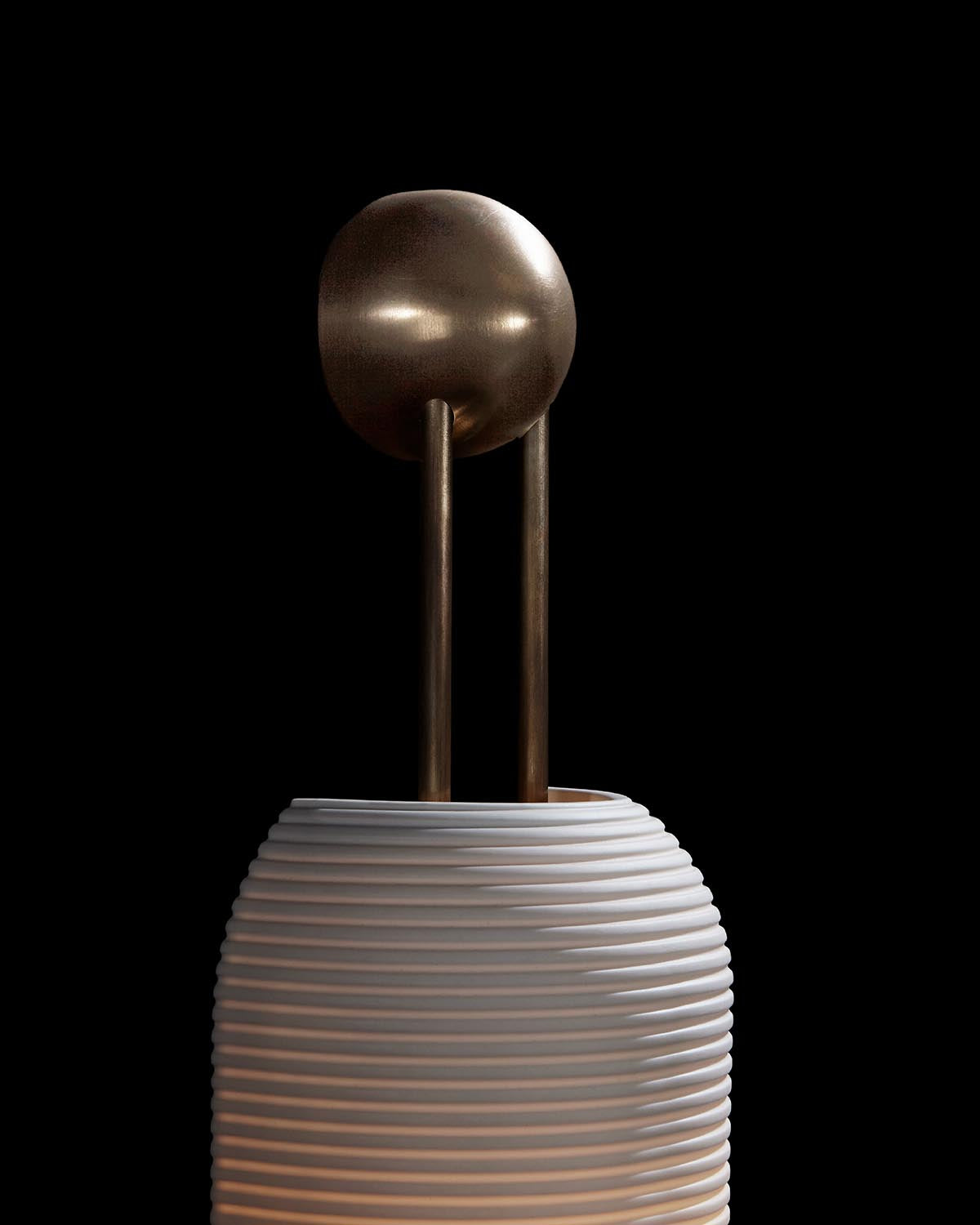 Wall sconce with ridged white cylindrical shade and aged brass spherical fixture with two vertical rods against black background