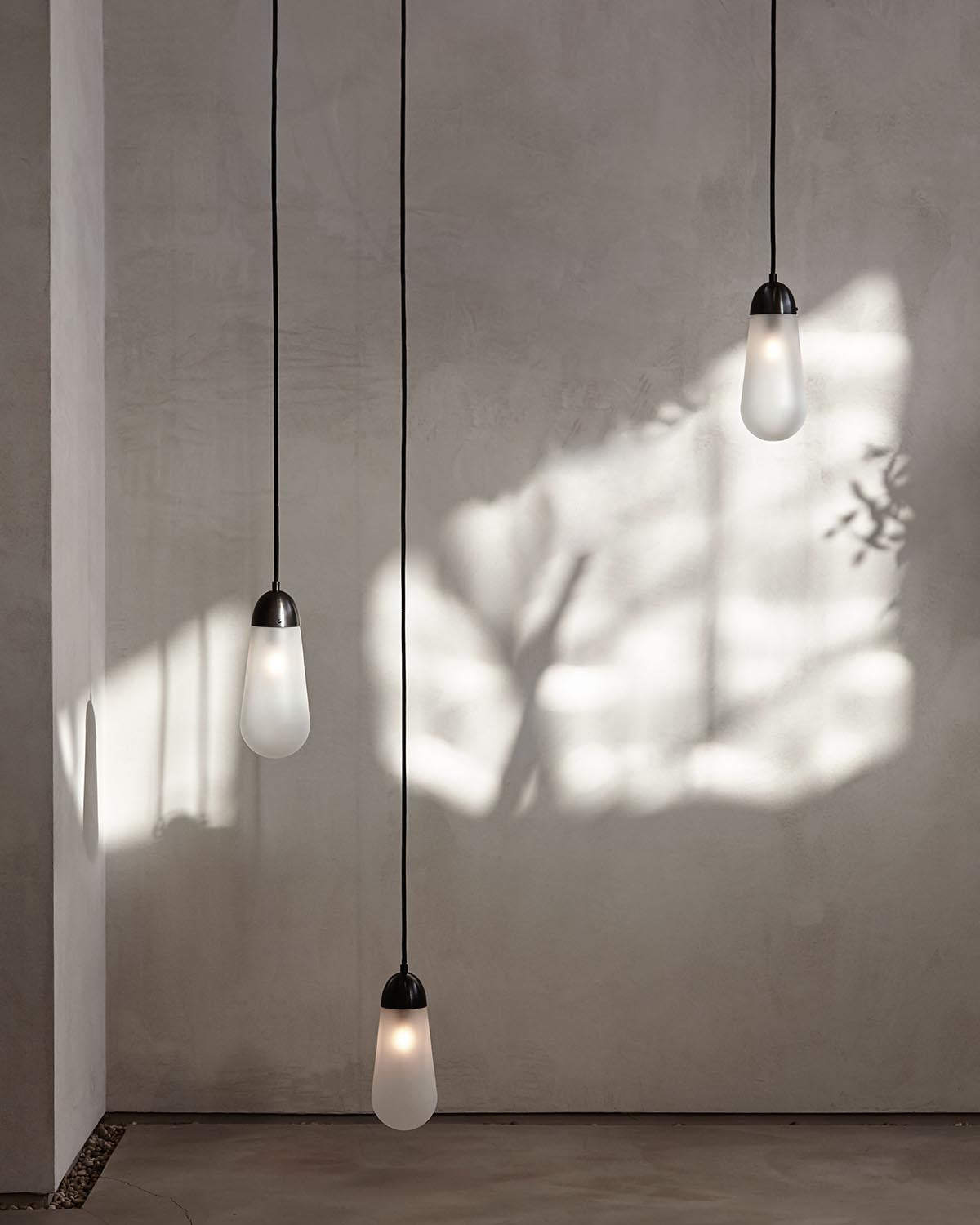 Three elongated frosted glass pendant lights hanging at different heights against a textured grey wall with soft shadows