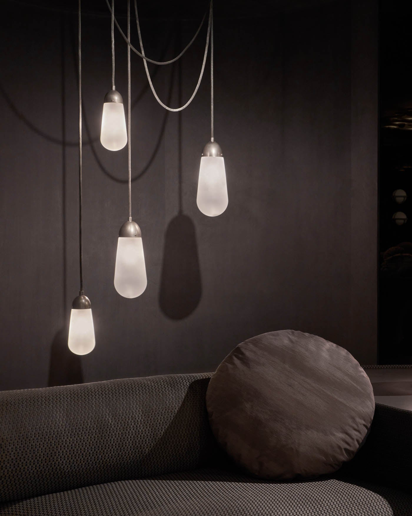 Four elongated frosted glass pendant lights with metal caps hanging over a textured dark sofa with a round cushion