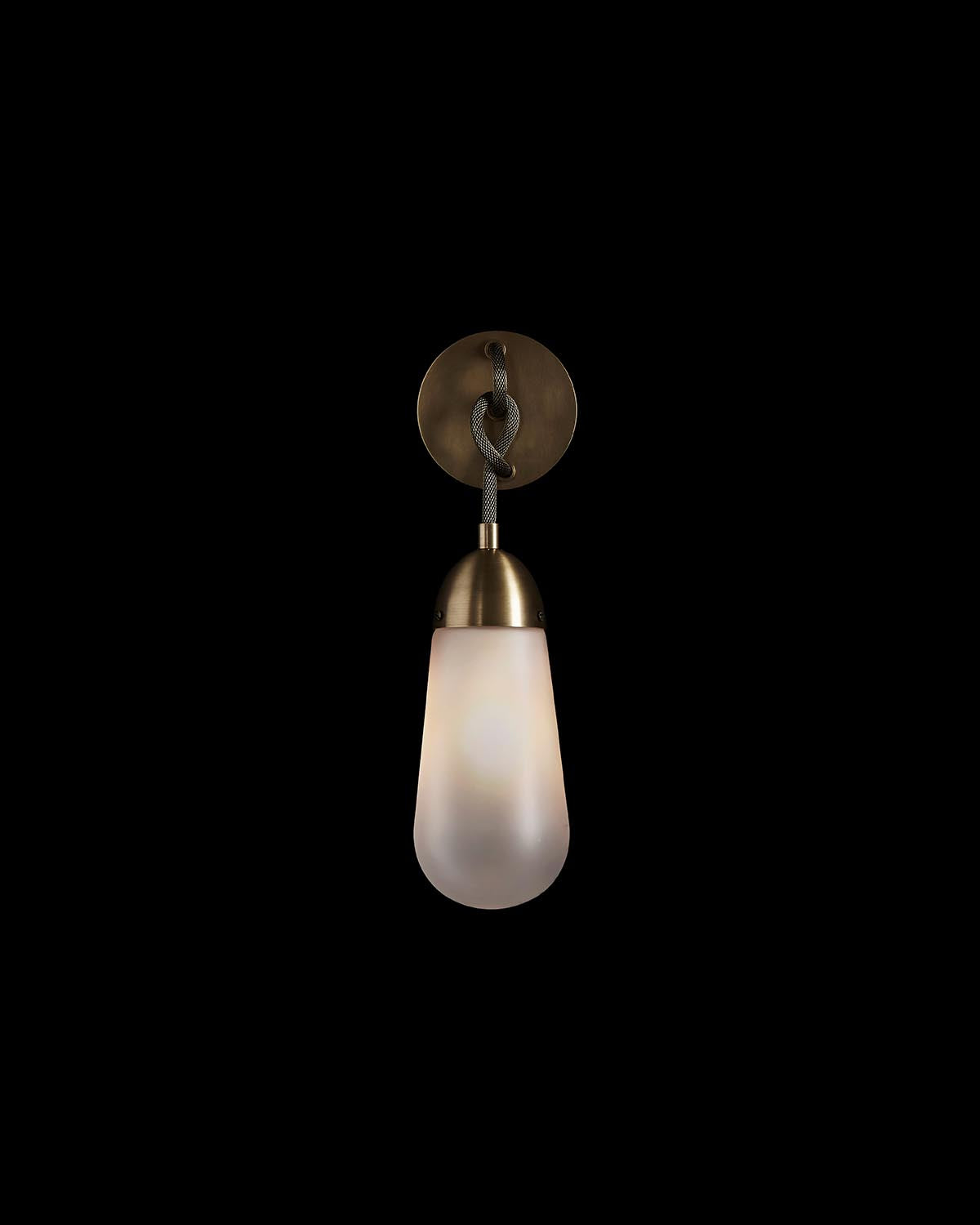 Wall sconce with frosted cylindrical glass shade hanging from knotted dark cord and brass circular wall plate