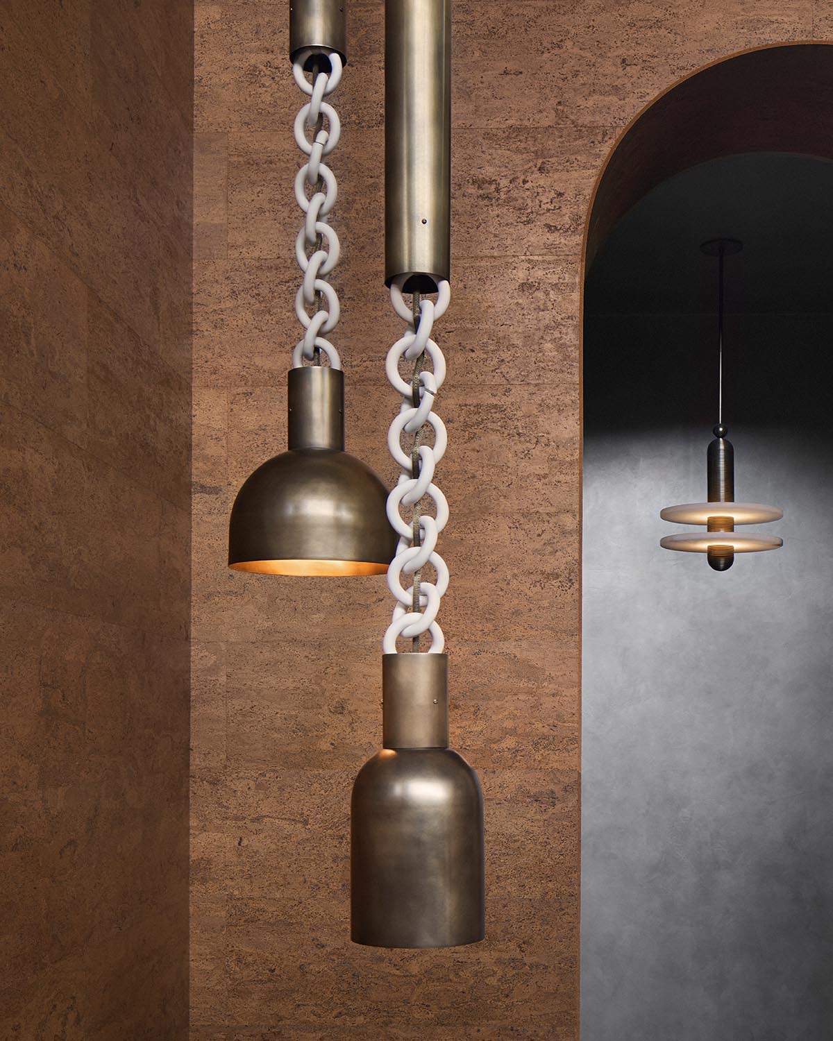 Two antique brass pendant lights suspended by white linked chains against a textured brown wall and arched doorway