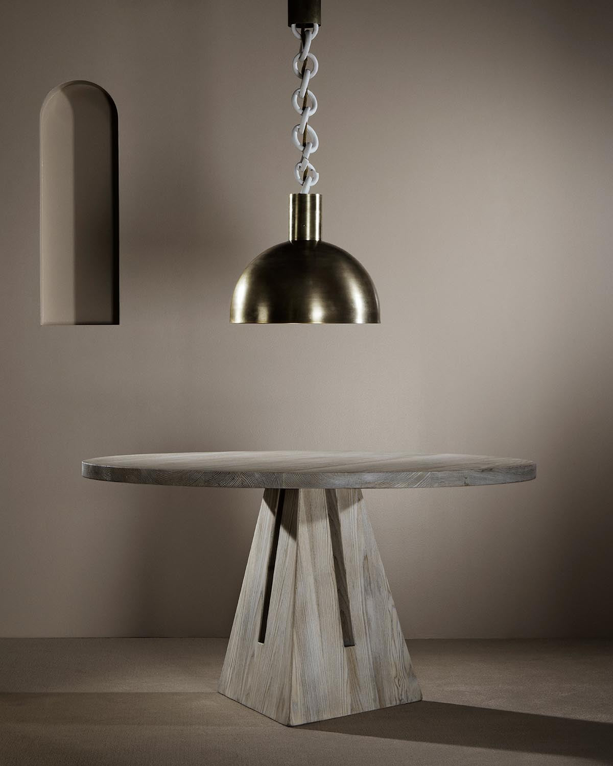 Brushed metal dome pendant light suspended by linked chain over round wooden table with geometric base against beige wall