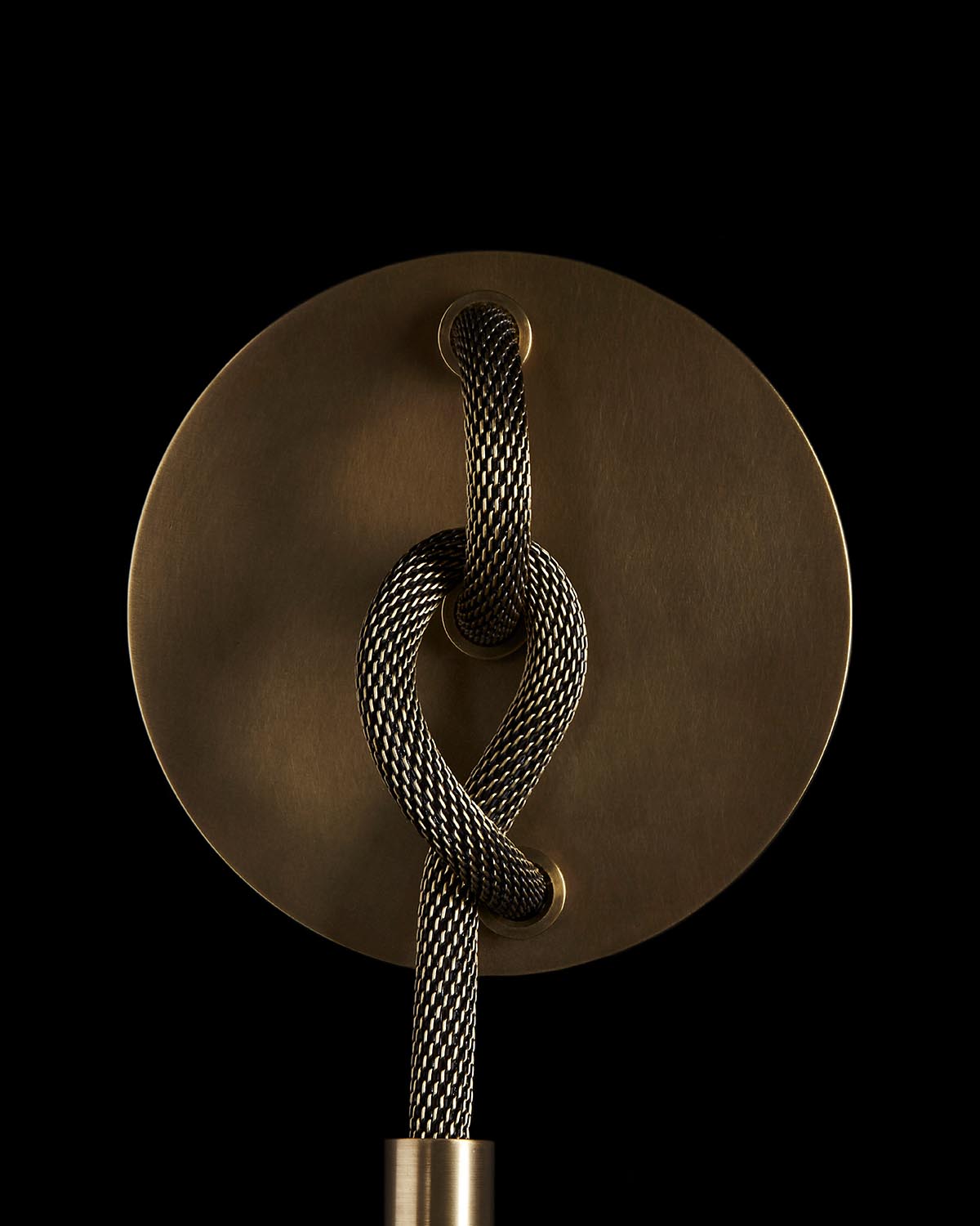 Close-up of a braided black and white cord looped through a round brushed brass wall plate against a black background