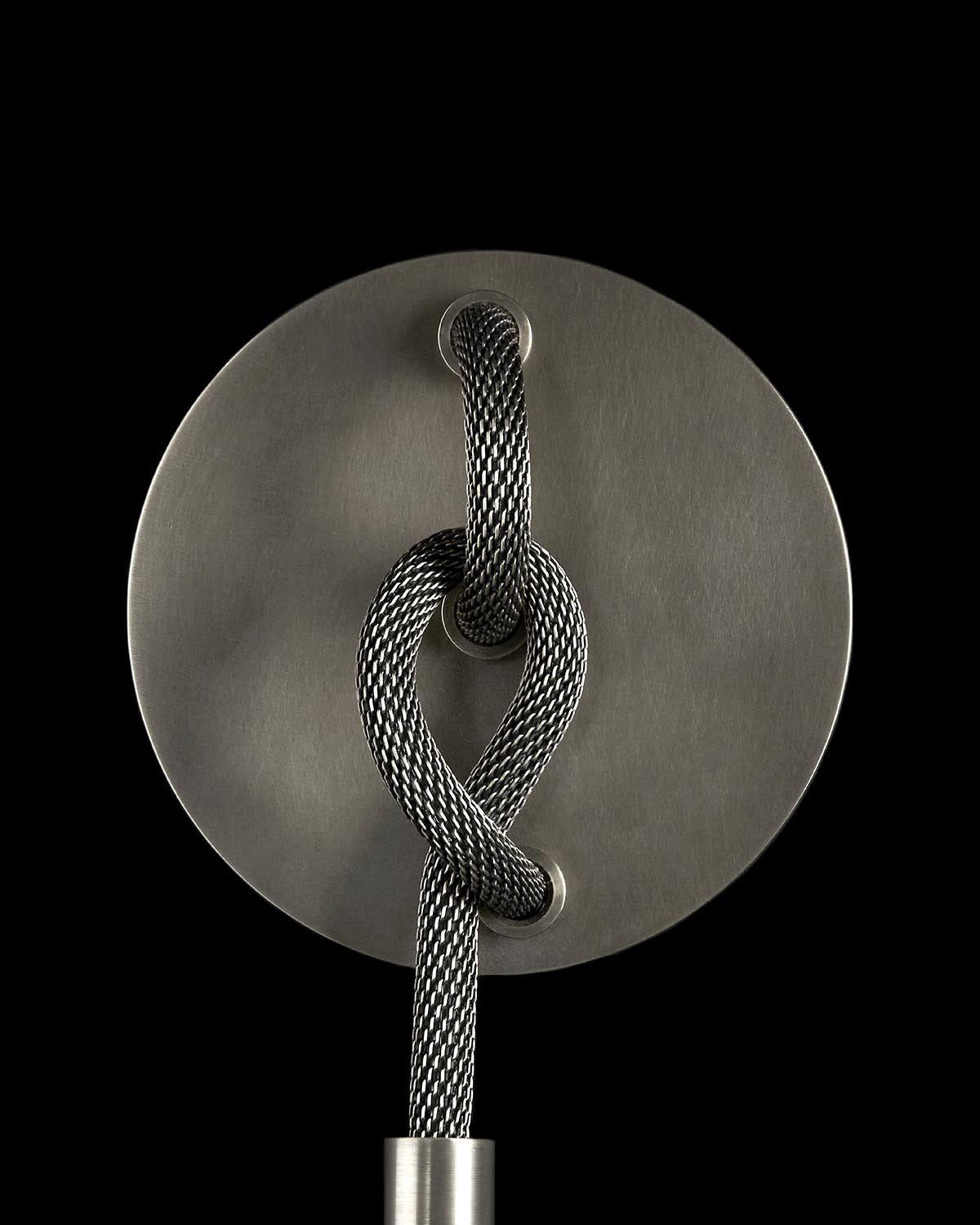 Close-up of a metallic braided cable threaded through a circular brushed metal plate against a black background
