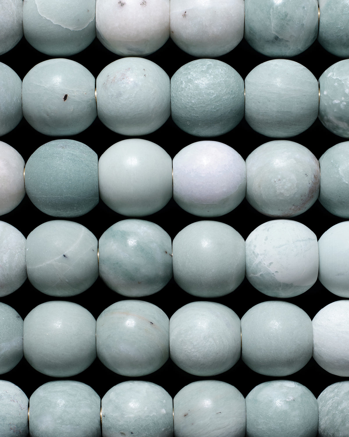 Close-up of polished round beads in soft green and pale blue hues arranged in horizontal rows on a black background