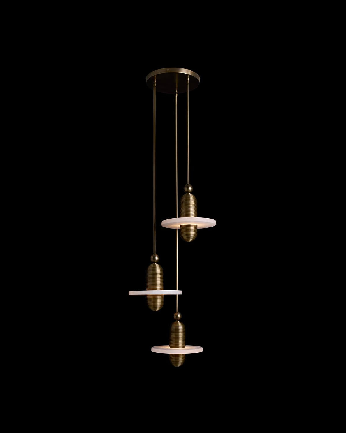 Three-tiered brass pendant light installation with cylindrical shades and glowing white discs against black background