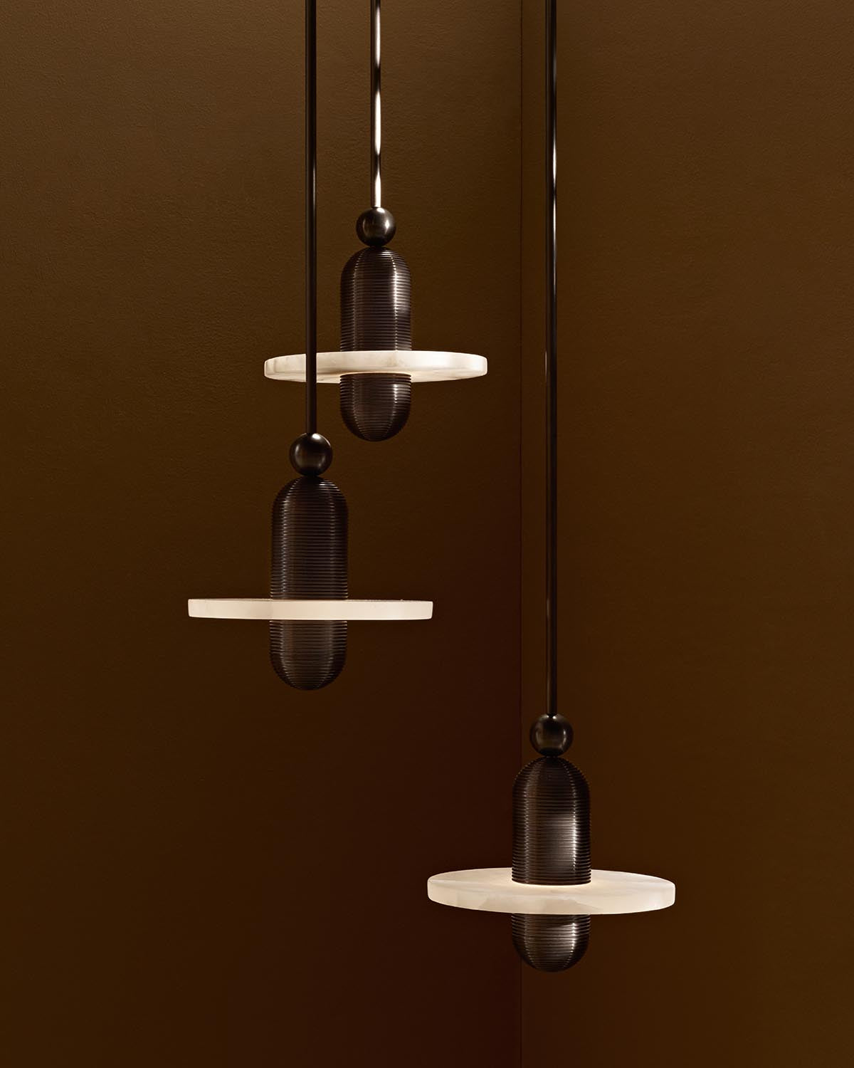 Three modern pendant lights with dark cylindrical bodies and translucent circular discs hanging against a brown background