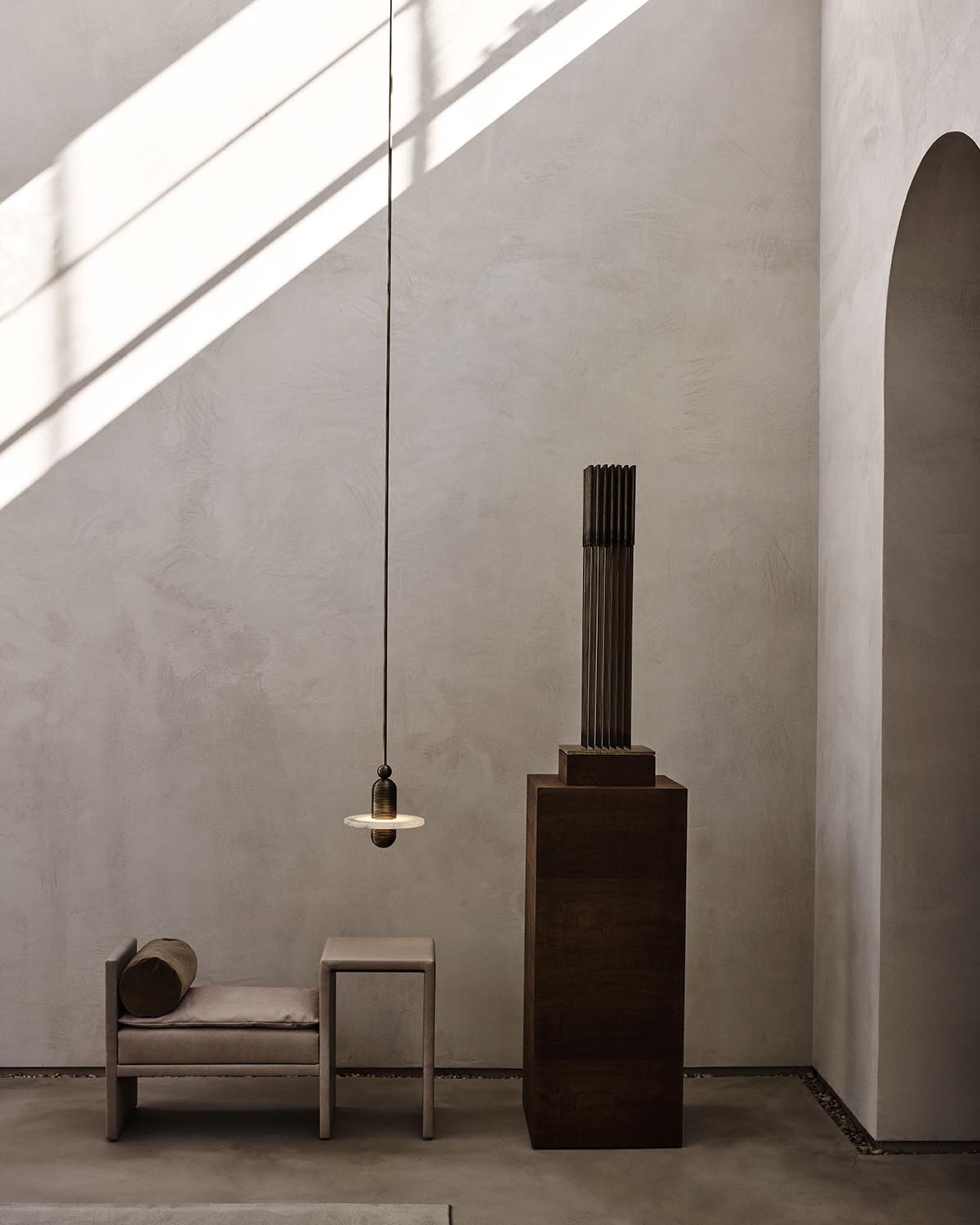 Minimalist interior featuring a textured beige wall, small wooden bench with cushion, table, and tall sculptural pedestal with light fixture above