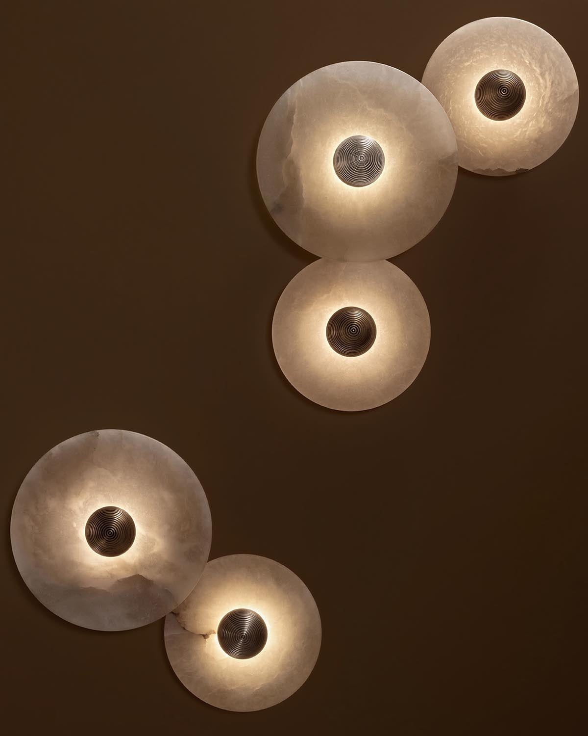 Two pairs of circular alabaster sconces with textured metal centers glowing softly on a brown wall background