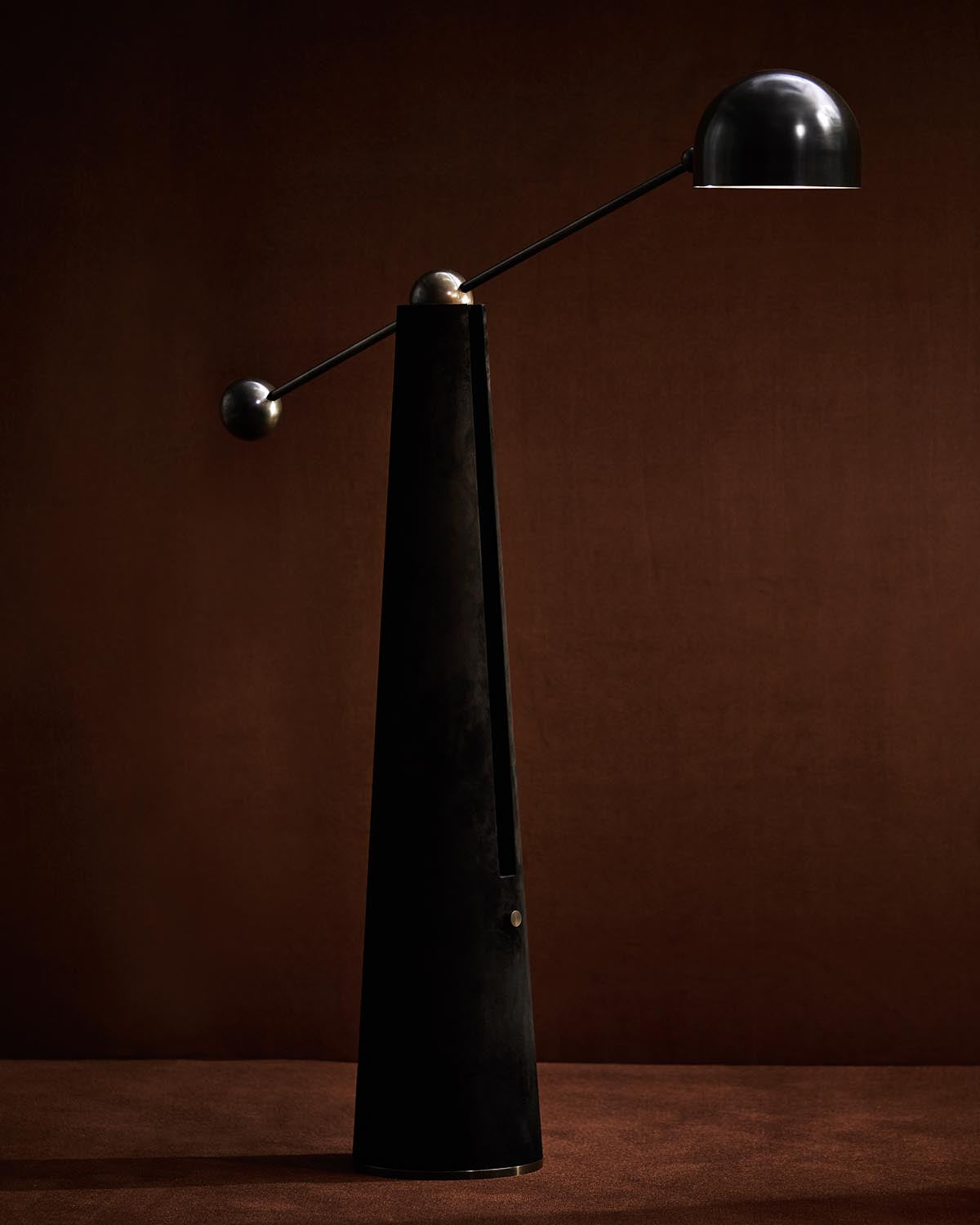 Tall black metronome reading lamp with a sleek dome-shaped shade and counterweight on a dark brown background