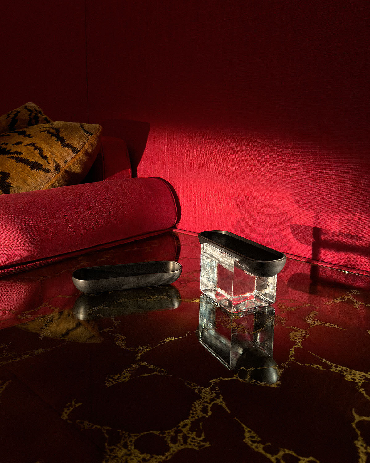 Two elongated black metal trays, one resting on a clear acrylic block, displayed on a glossy marble surface with red fabric backdrop