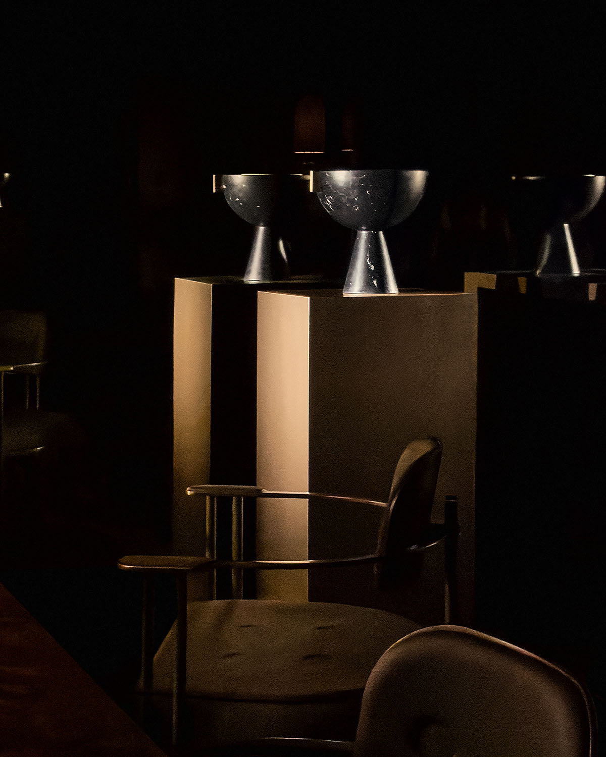 Metallic vessel with a rounded bowl shape on a pedestal stand displayed on a beige rectangular plinth in dim lighting