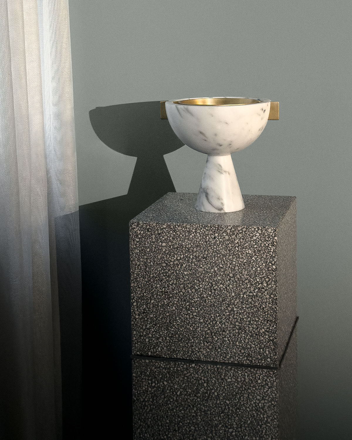 Marble vessel with brass handles on a textured dark stone pedestal against a muted green wall