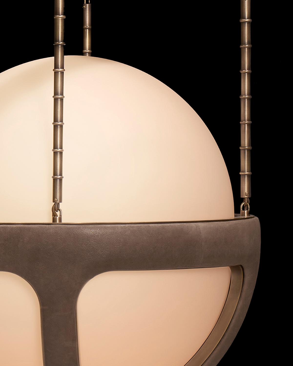 Close-up of a spherical pendant light with frosted glass and bronze metal frame suspended by textured rods against black background
