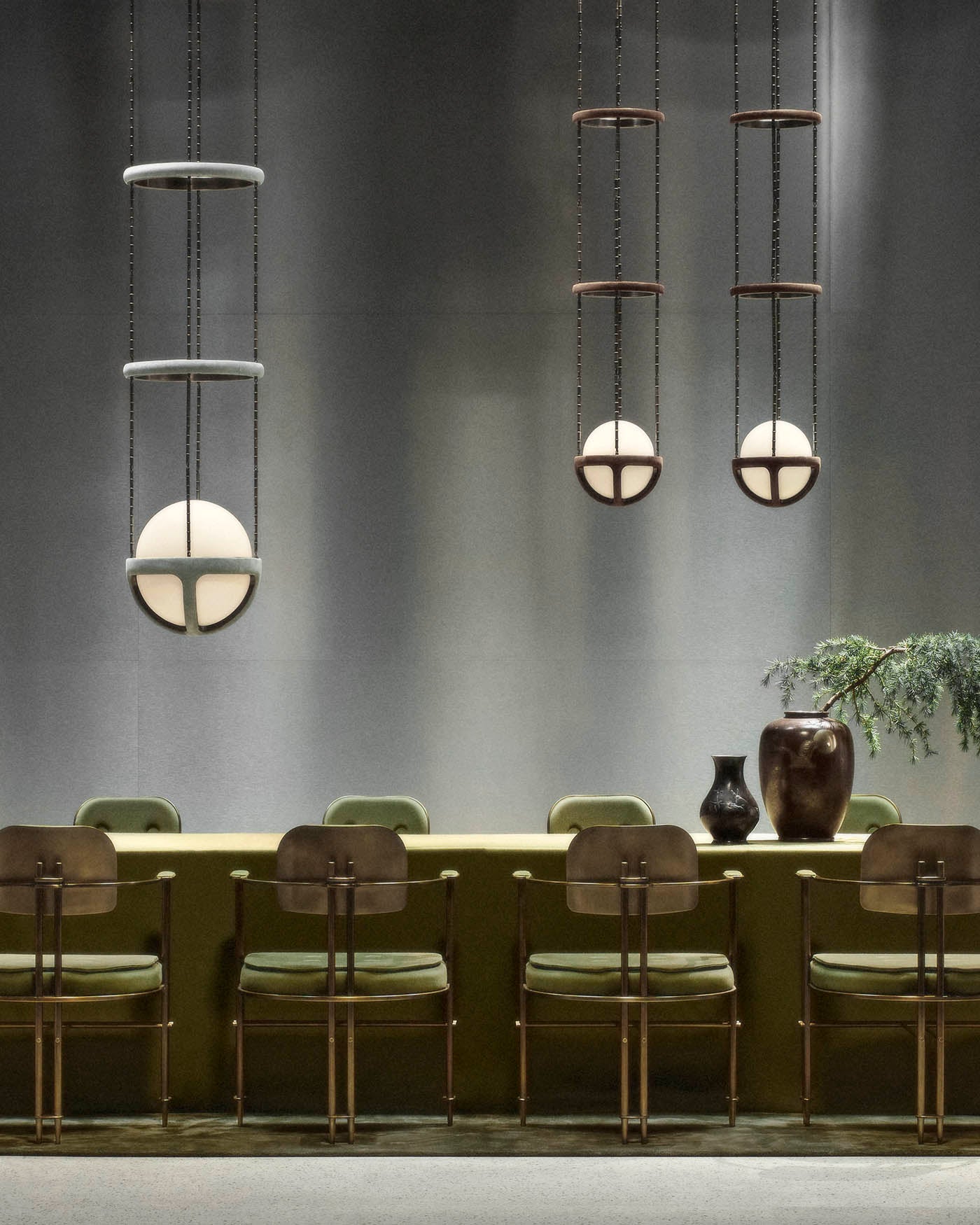 Three pendant lights with spherical bulbs suspended by metal frames above an olive green cushioned bench and vintage chairs