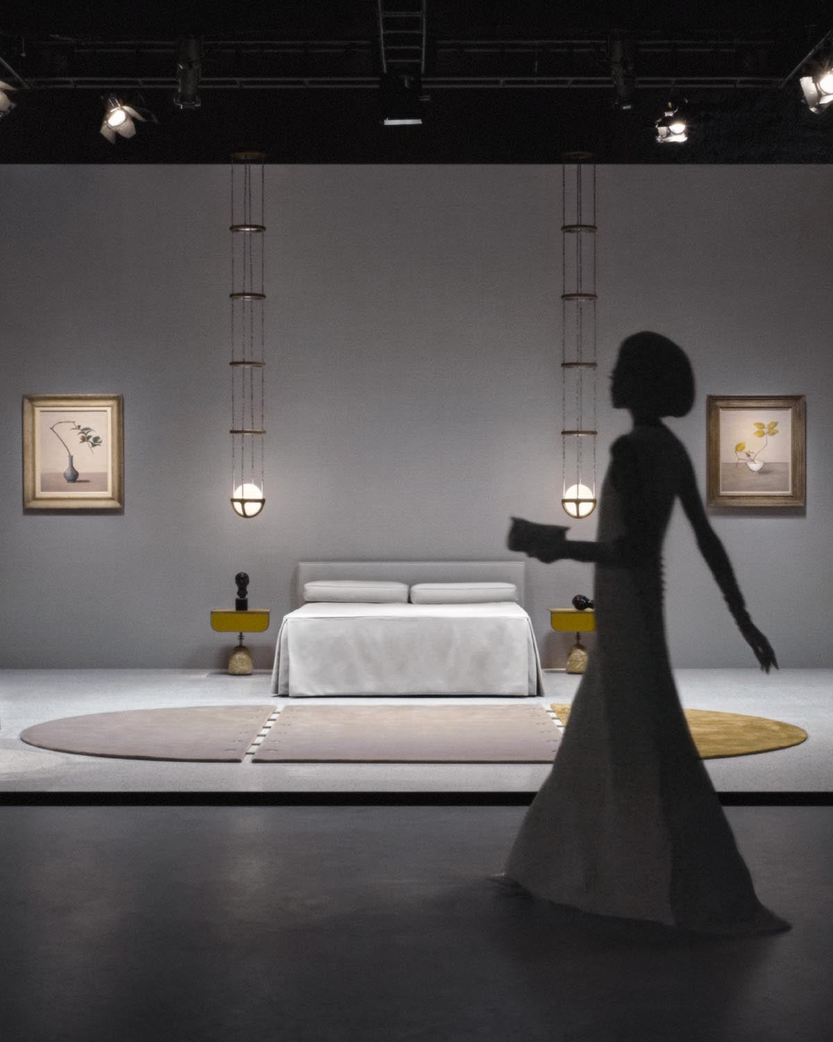 Minimalist bedroom scene with white bed, round pendant lights suspended by vertical metal rods, and framed botanical art on gray wall