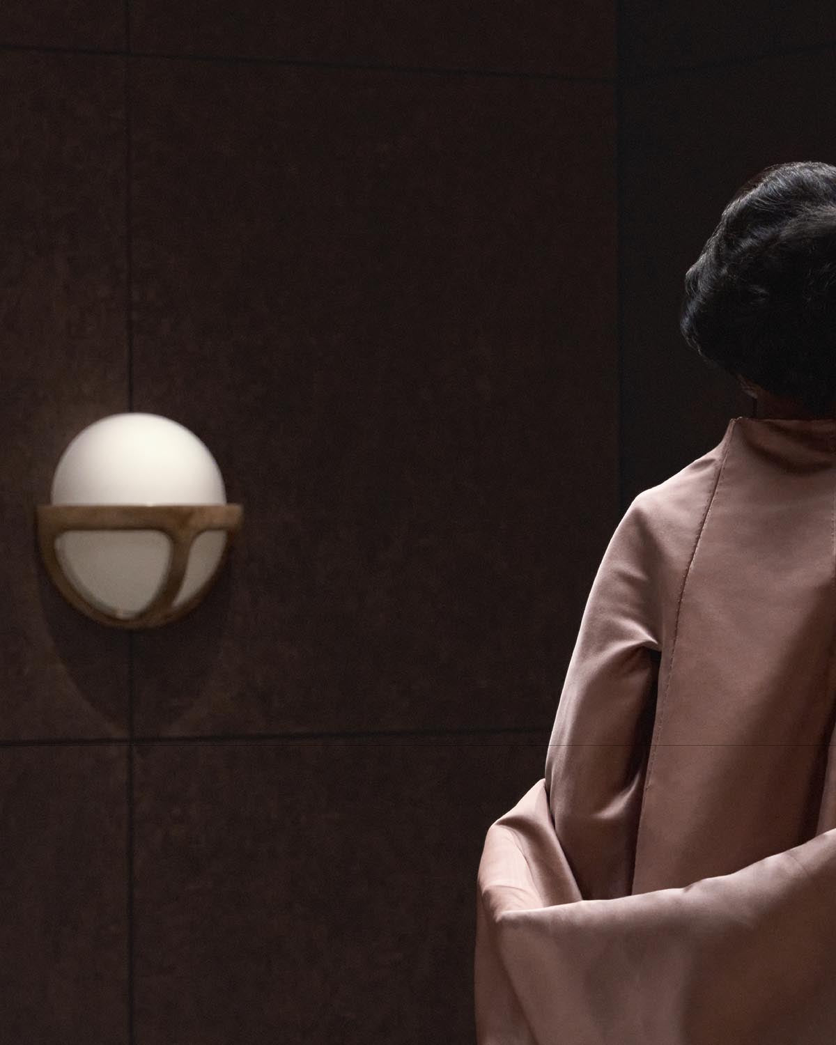 Wall-mounted spherical white glass sconce with curved wooden bracket on dark textured wall, partially obscured by person in pale pink garment