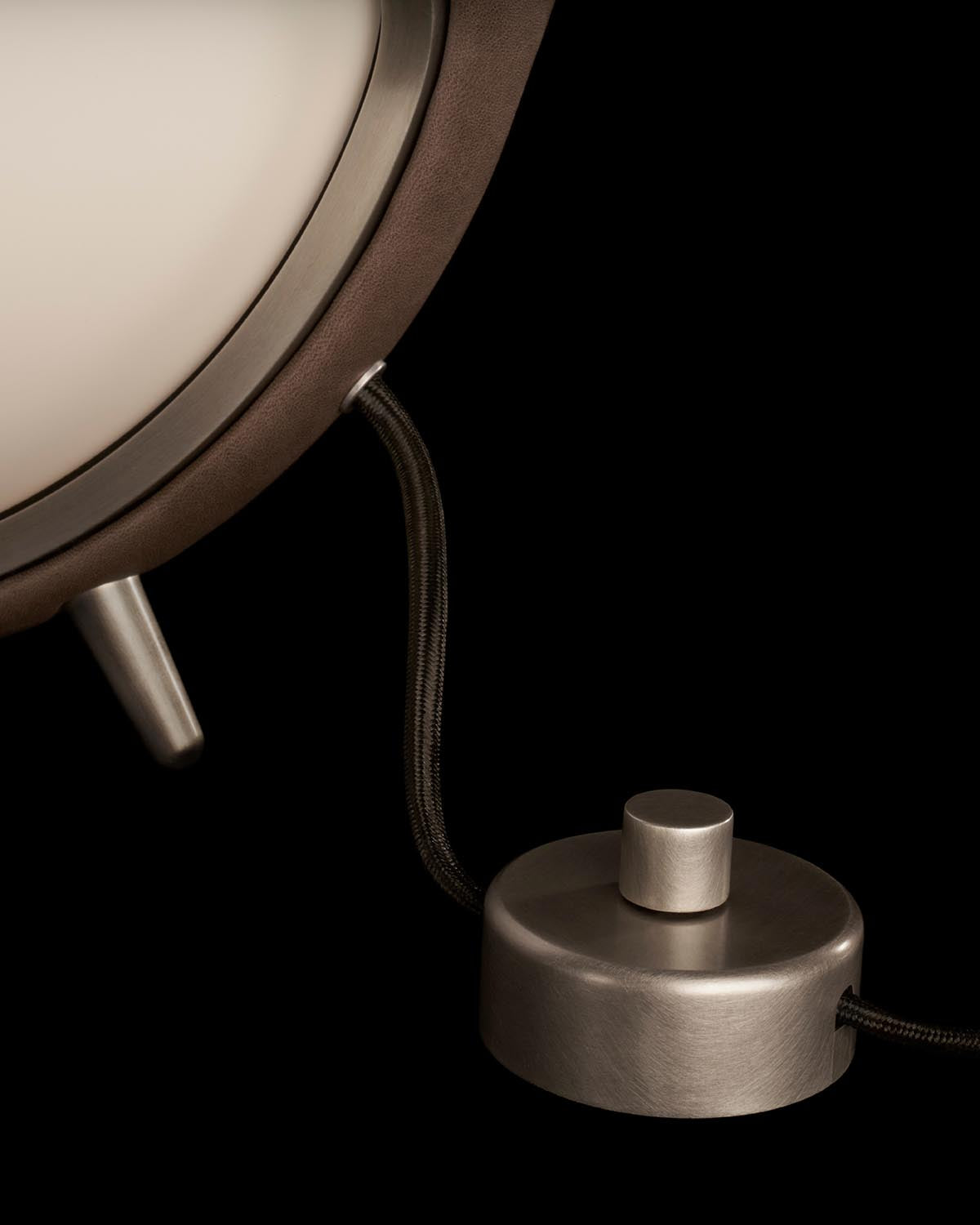 Close-up of a spherical table lamp with brushed metal base and black fabric-covered cord against a black background