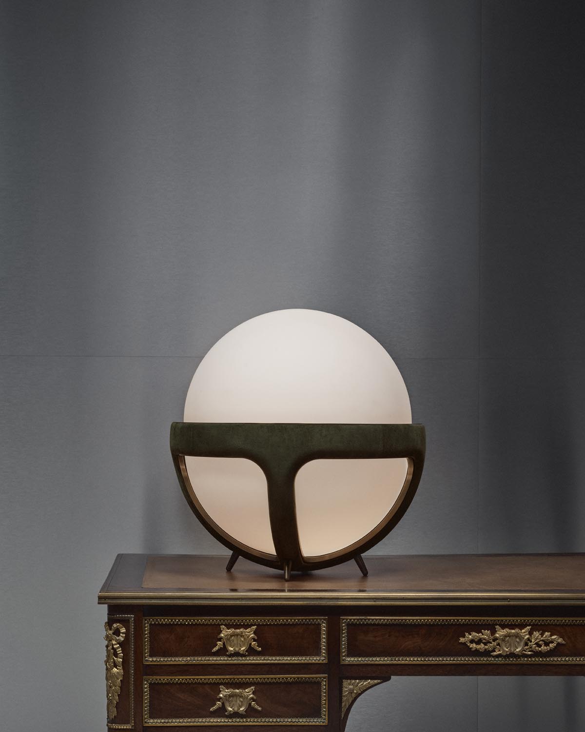 Round white glass table lamp with dark curved metal frame on vintage wooden desk against gray wall background