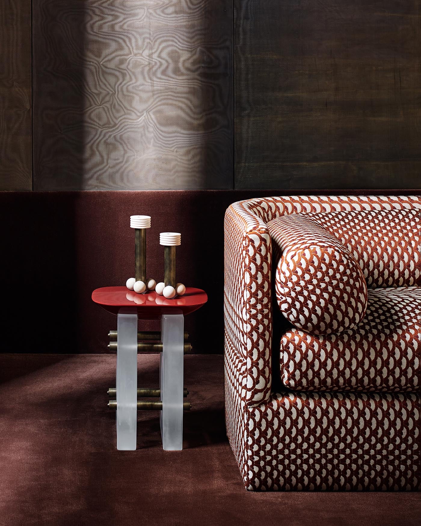 Small cocktail table with red top and clear legs next to a patterned curved sofa on a dark brown carpet background