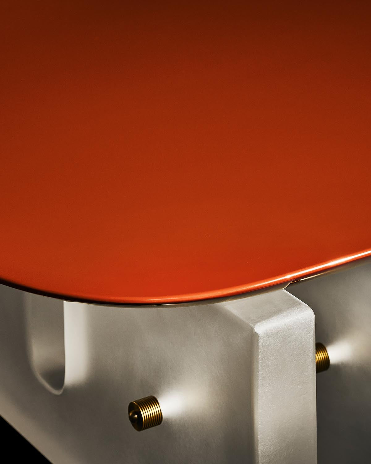 Close-up of a coffee table with smooth matte red top and concrete base secured by brass bolts on a black background