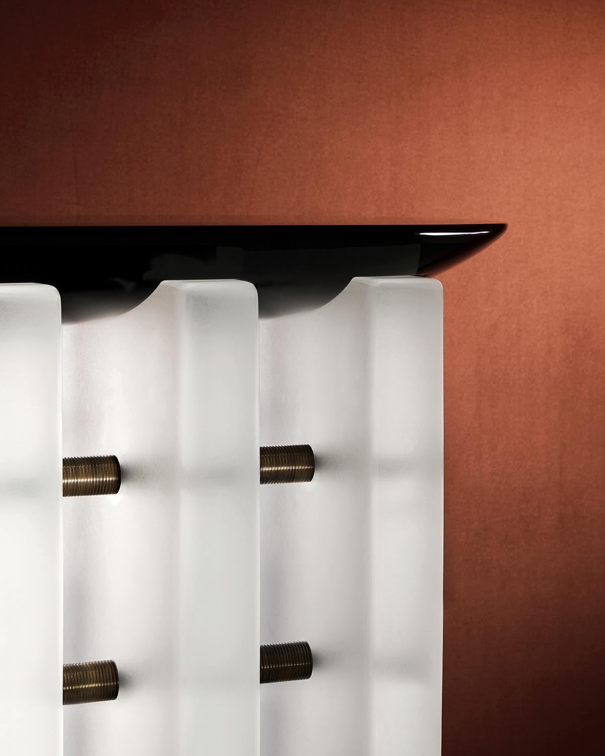Close-up of a minimalist side table featuring frosted glass panels connected by brass rods and a sleek black top against a brown background
