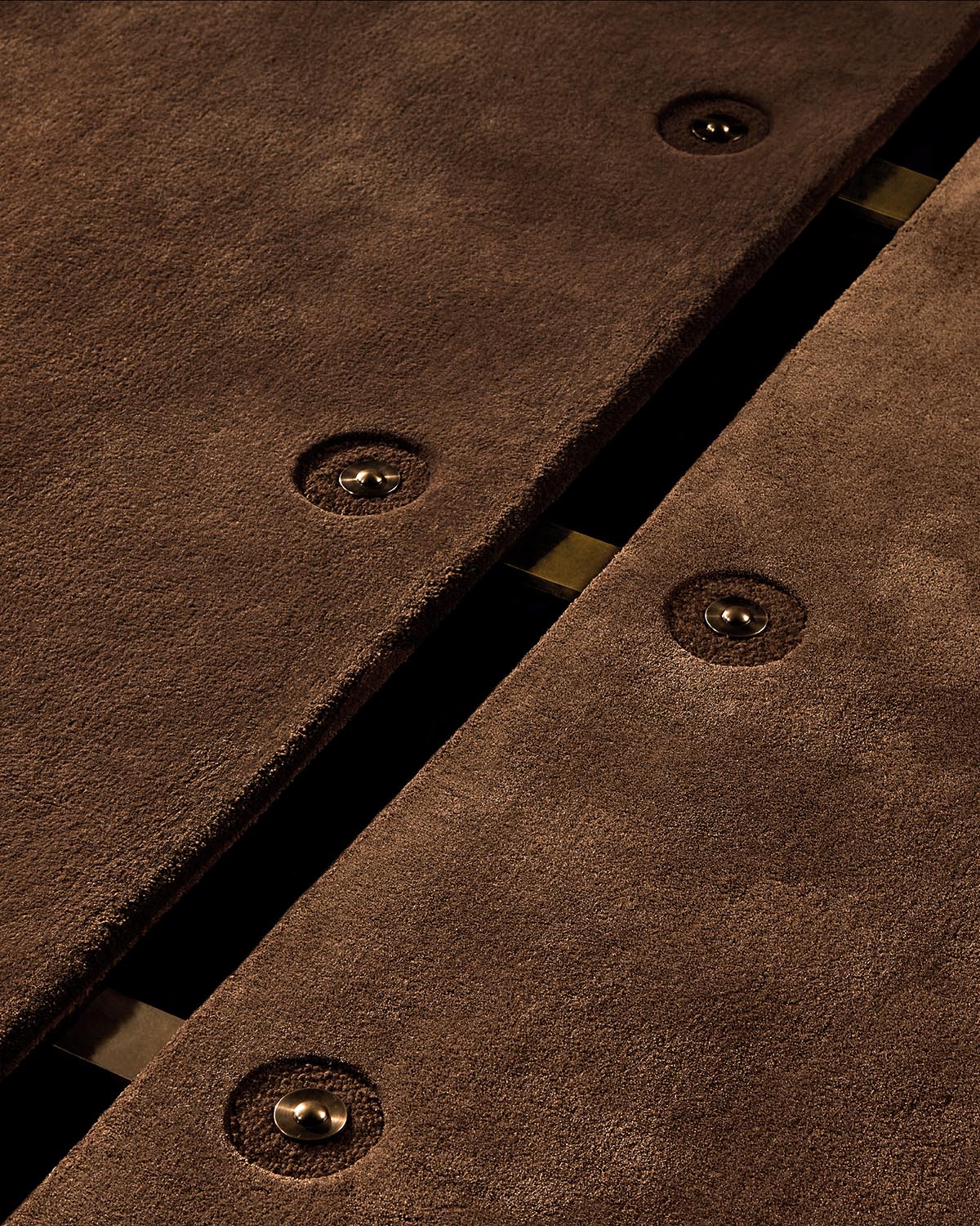 Close-up of brown suede panels with circular metal fasteners and a glimpse of brass hardware underneath against dark background