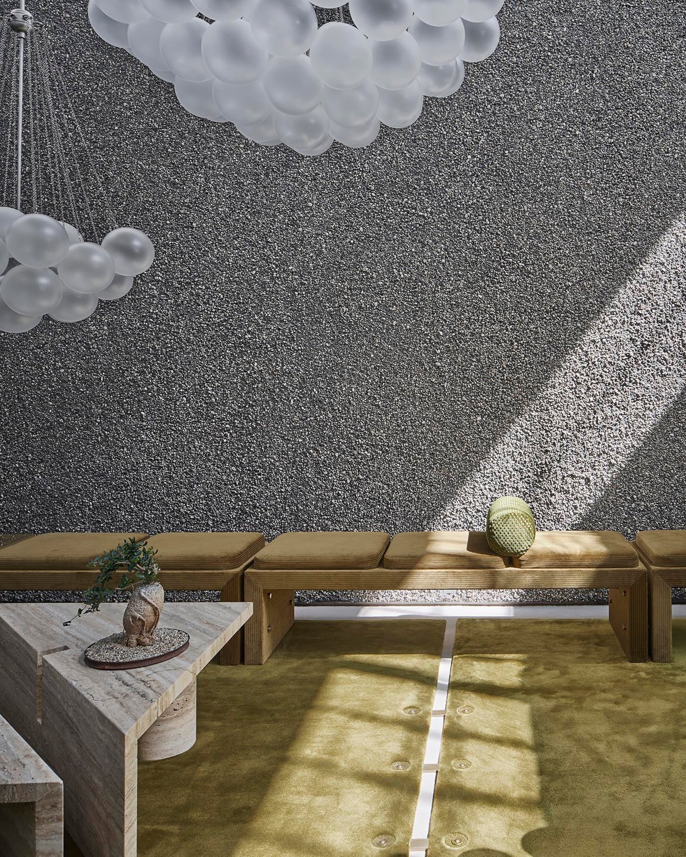 Modern seating area with long cushioned benches, textured gray wall, geometric stone table, and cluster pendant lights