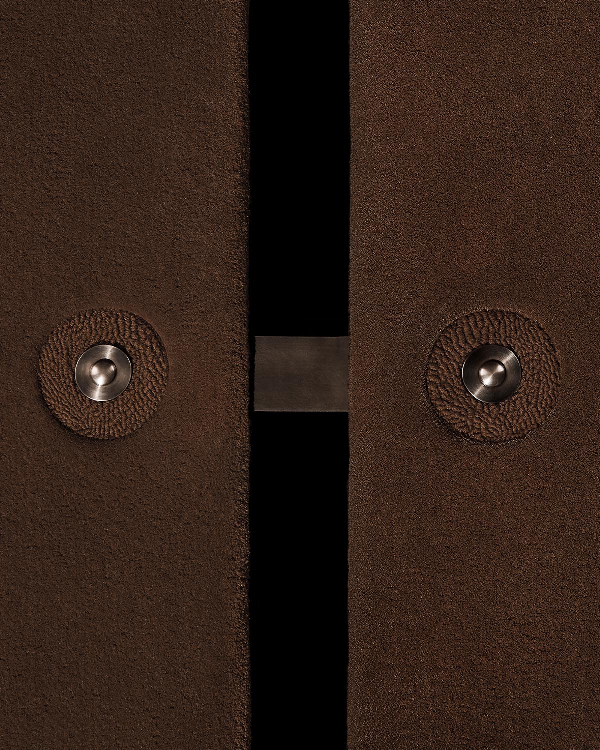 Close-up of textured brown fabric panels with circular metal hardware and a central dark rectangular connector against black background